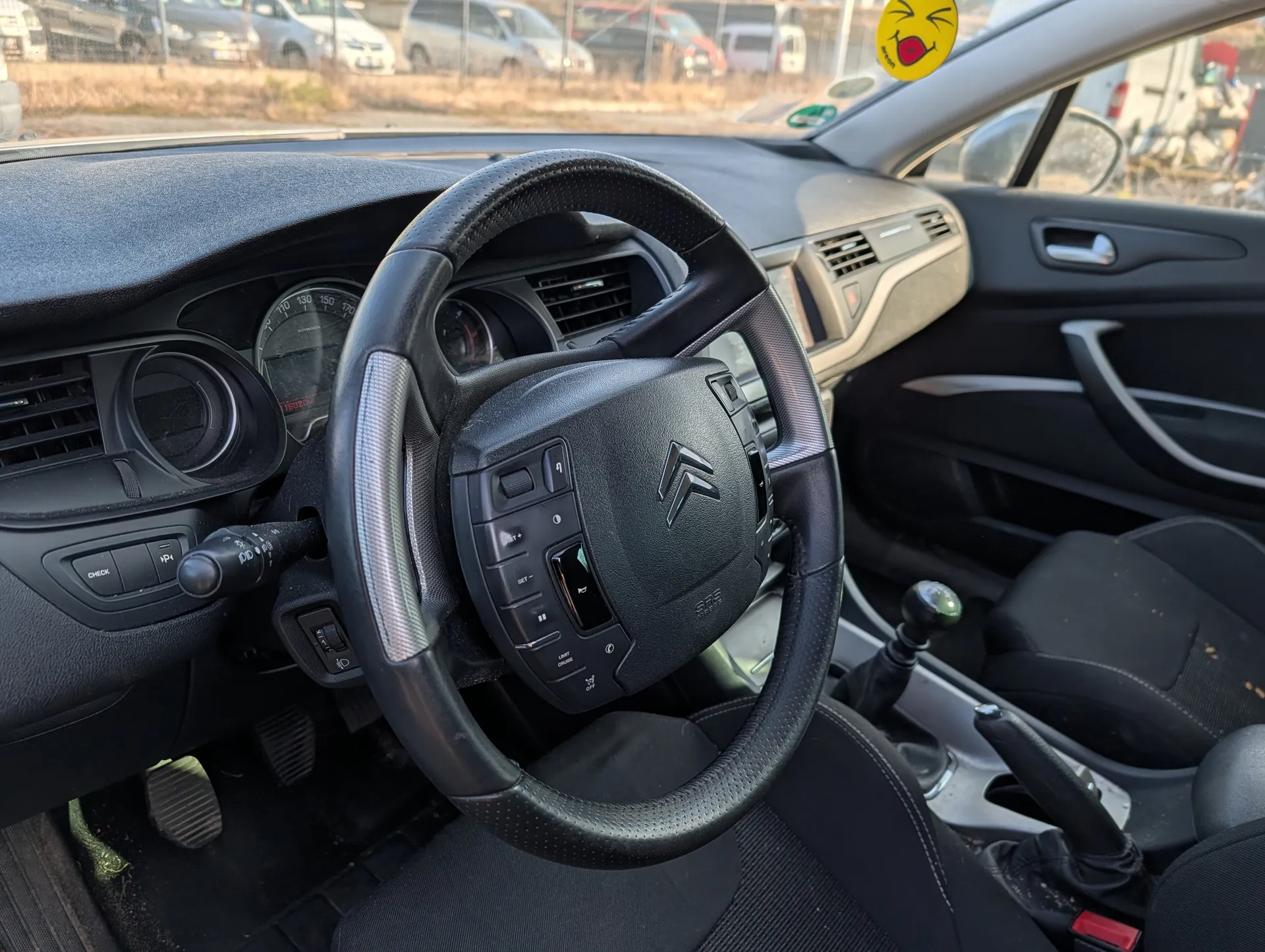 Wnętrze samochodu Citroën z widocznym kierownicą z logo marki, czarną deską rozdzielczą z licznikami prędkości i obrotomierzem, przyciskami sterującymi oraz dźwignią zmiany biegów. Na desce rozdzielczej widoczne są przyciski oznaczone napisami 'CHECK' i 'IP'. W tle przez przednią szybę widać zaparkowane samochody i suchą trawę. Na szybie umieszczony jest żółty naklejka z uśmiechniętą twarzą. Siedzenia są czarne, a podłoga pokryta jest brudnym dywanikiem. W prawym dolnym rogu widoczna jest czerwona klamra pasa bezpieczeństwa.