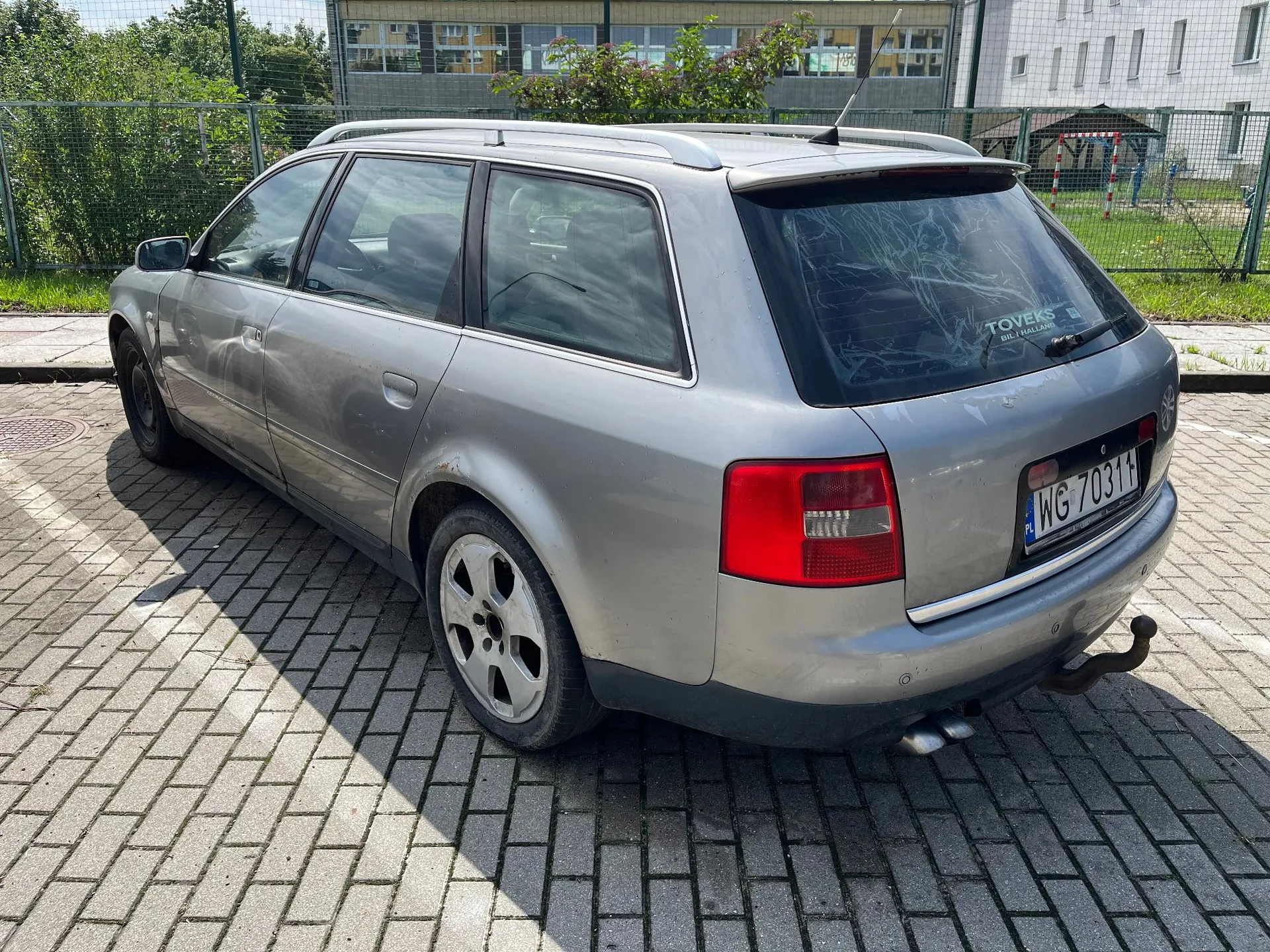 Srebrny samochód typu kombi zaparkowany na kostce brukowej. Pojazd jest widoczny z tyłu i z boku. Na tylnej klapie znajduje się tablica rejestracyjna o numerze WG 70311. Samochód ma hak holowniczy oraz widoczne uszkodzenia lakieru i rdzę na nadkolu. W tle widać zielone krzewy, ogrodzenie oraz biały budynek.