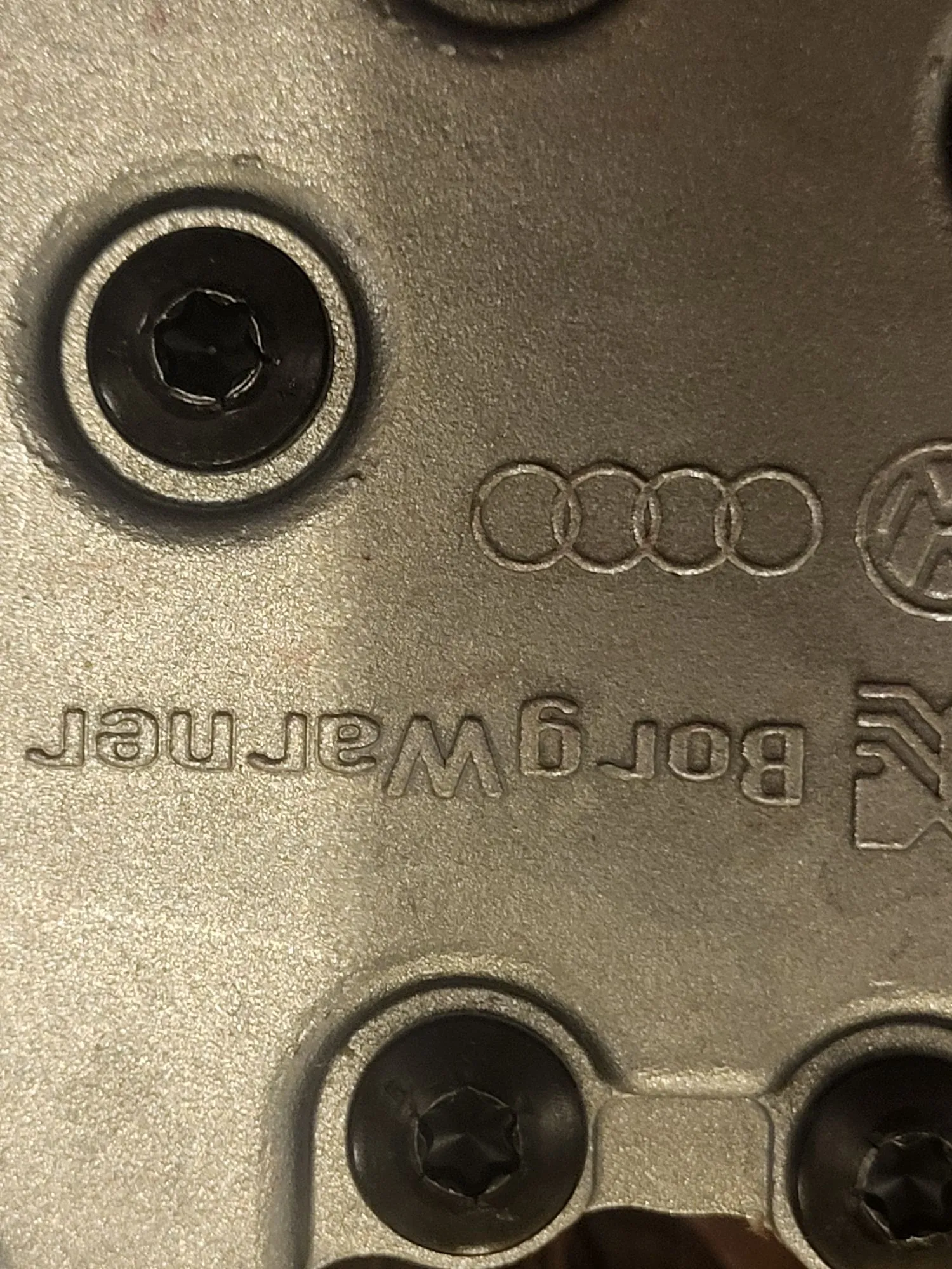 Zbliżenie na metalową, srebrną część samochodową z grawerowanym napisem „BorgWarner” oraz logo Audi składającym się z czterech przeplatających się pierścieni. Widoczne są dwie czarne śruby z łbem imbusowym.