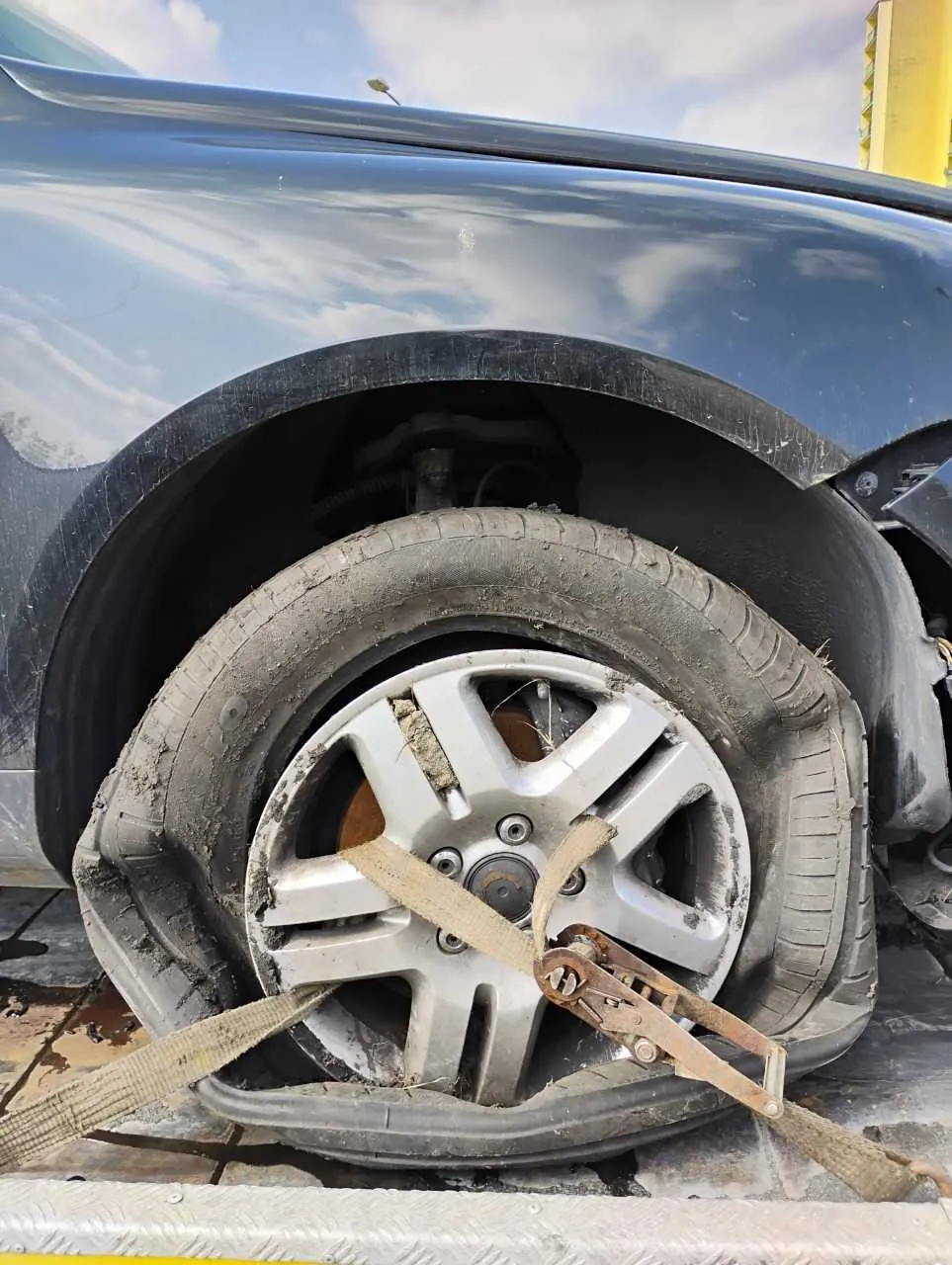 Zbliżenie na uszkodzone koło samochodu ciemnoniebieskiego koloru. Opona jest zdeformowana i zabrudzona, a srebrna felga z pięcioma promieniami ma widoczne ślady brudu. Koło jest przywiązane taśmą z metalowym zapięciem do platformy załadunkowej. W tle widoczne niebo z chmurami oraz fragment żółtej konstrukcji. Samochód znajduje się na metalowej powierzchni z widocznymi plamami.