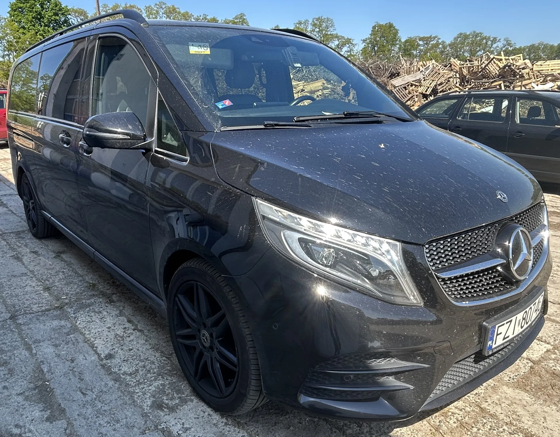 Czarny samochód Mercedes-Benz Vito zaparkowany na brukowanej powierzchni. Widoczna tablica rejestracyjna FZ1 8074, logo Mercedes-Benz na atrapie chłodnicy, czarne felgi i lusterko boczne. W tle drugi ciemny samochód, stosy drewnianych palet oraz drzewa pod jasnym niebem. Na przedniej szybie naklejki, w tym żółta z napisem 10.