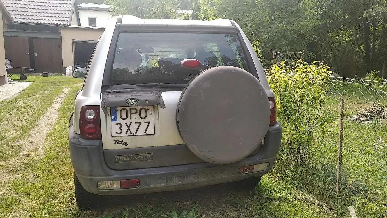 Tylny widok srebrnego samochodu terenowego Land Rover Freelander z zamontowanym kołem zapasowym na klapie bagażnika. Pojazd posiada polską tablicę rejestracyjną o numerze OPO 3X77. Na klapie widoczne są napisy 'Td4' oraz 'FREELANDER'. Samochód stoi na trawiastym podjeździe przed domem z garażem, w otoczeniu zieleni i drzew.