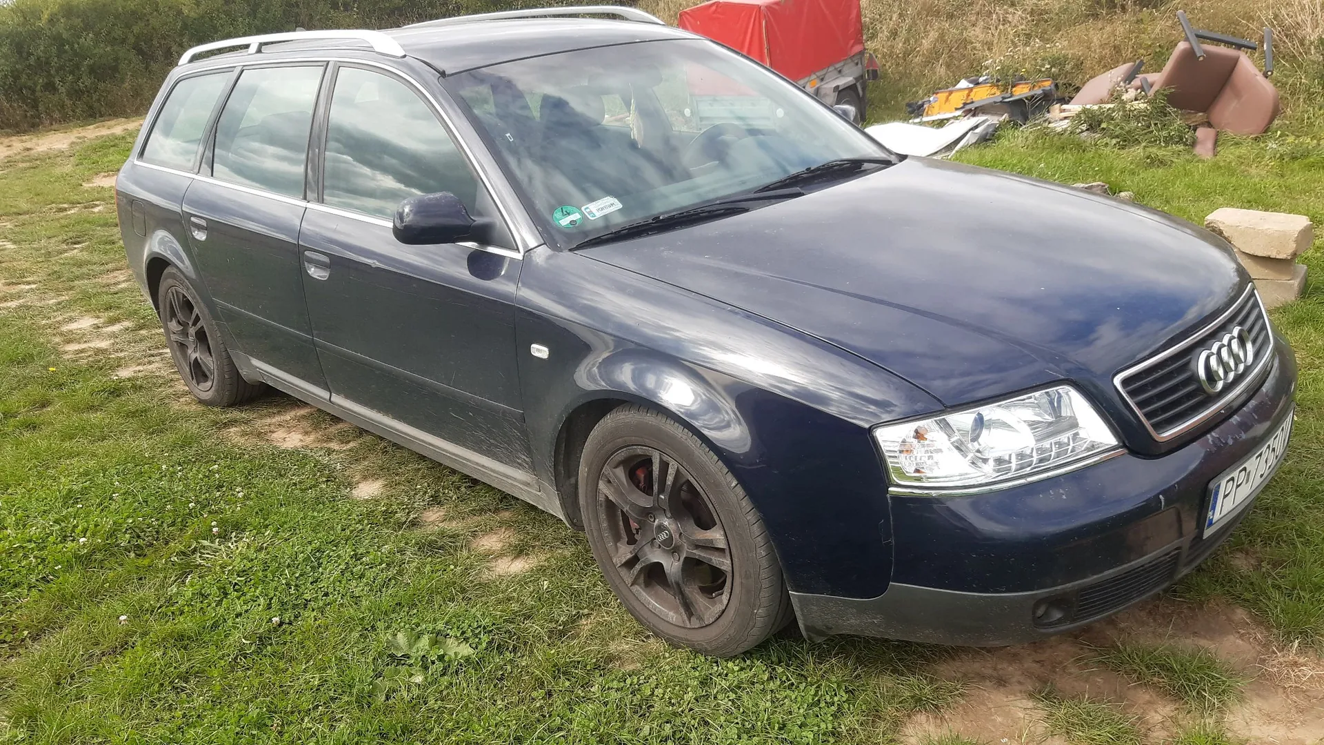 Ciemnoniebieski samochód marki Audi w nadwoziu kombi, zaparkowany na trawiastym terenie. Pojazd jest widoczny z przodu i z boku. Na masce i karoserii widoczne są zabrudzenia. W tle znajduje się zielona trawa, krzewy oraz fragmenty sprzętów i czerwona plandeka.