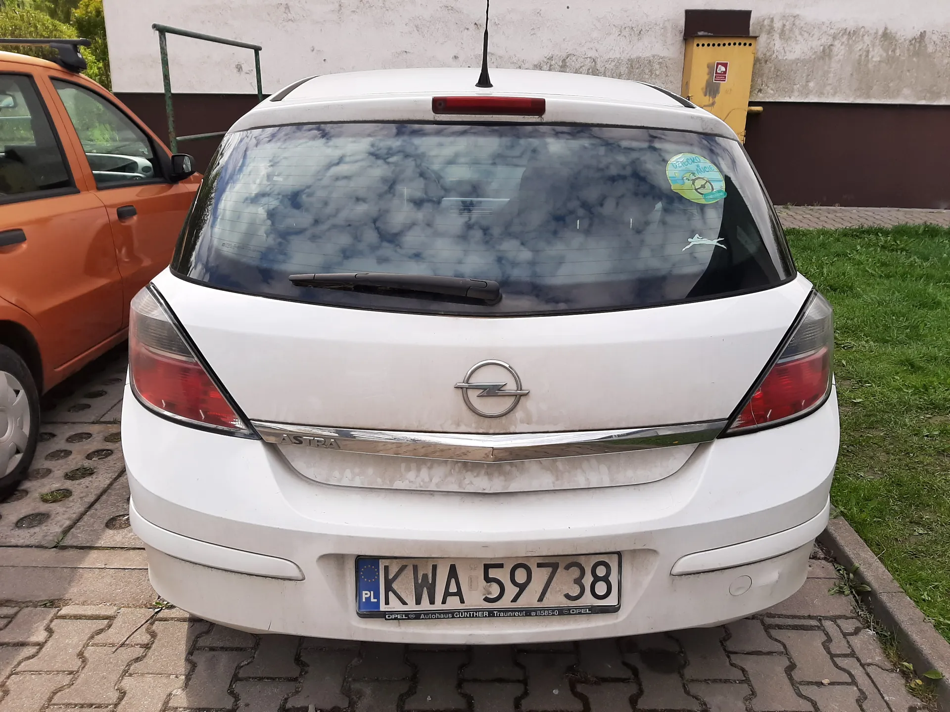 Tył białego samochodu Opel Astra z tablicą rejestracyjną PL KWA 59738. Na szybie tylnej naklejka z kreskówką i biały symbol. Po lewej stronie pomarańczowy samochód, na tle budynek z białą i brązową ścianą oraz żółty pojemnik. Samochód zaparkowany na brukowanej powierzchni obok trawnika.