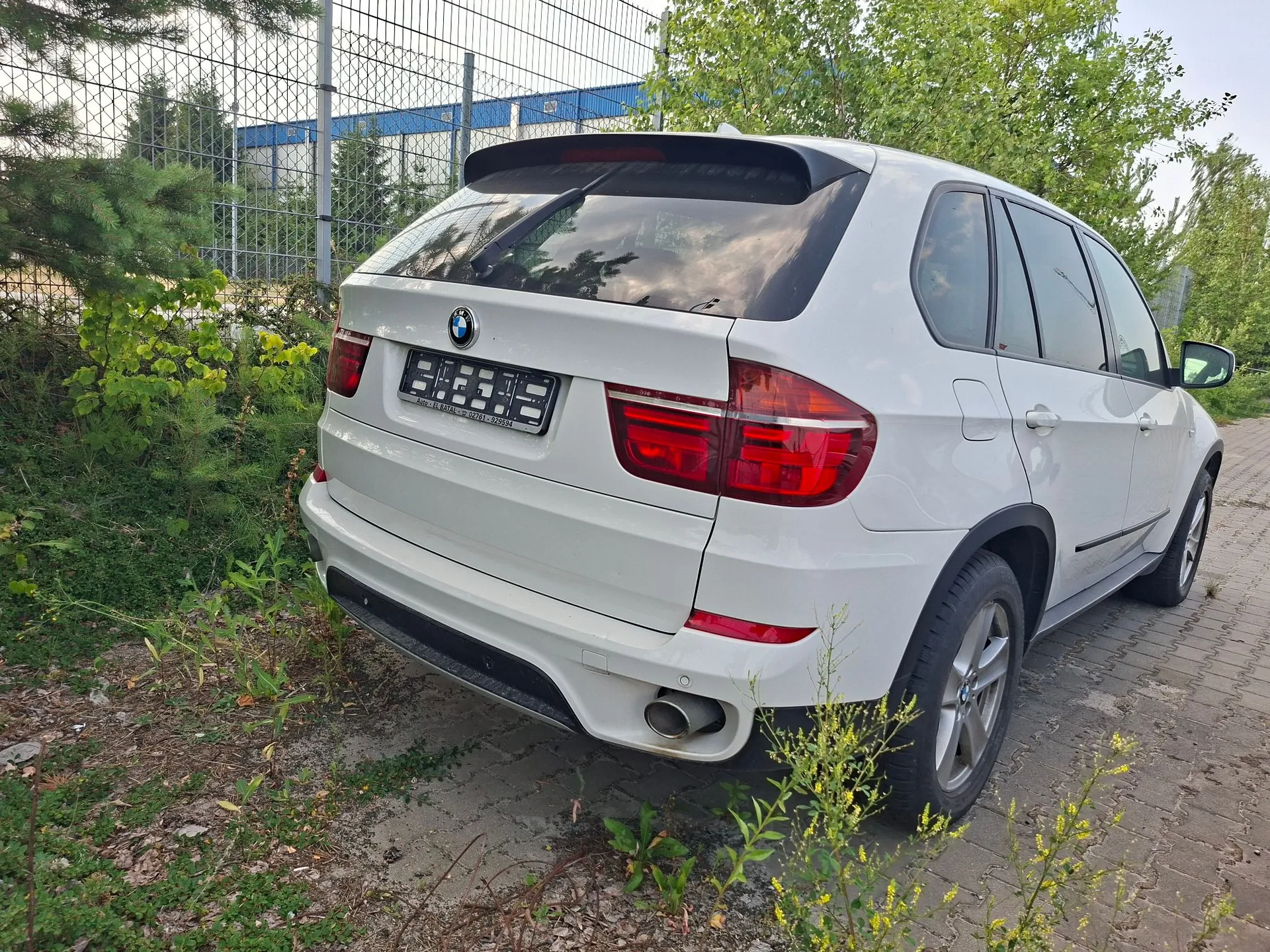 Tylny widok białego samochodu marki BMW typu SUV, zaparkowanego na kostce brukowej obok zieleni i metalowego ogrodzenia. Pojazd posiada czarny spojler nad tylną szybą, ciemne szyby oraz widoczną rurę wydechową po prawej stronie. W miejscu tablicy rejestracyjnej znajduje się czarna ramka z białymi napisami.