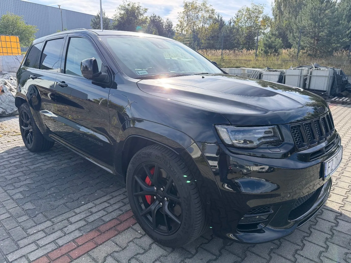 Czarny samochód marki Jeep Grand Cherokee zaparkowany na kostce brukowej. Pojazd jest widoczny z przodu i z boku, ma czarne felgi z czerwonymi zaciskami hamulcowymi. W tle widać budynek przemysłowy, drzewa oraz stosy białych palet.