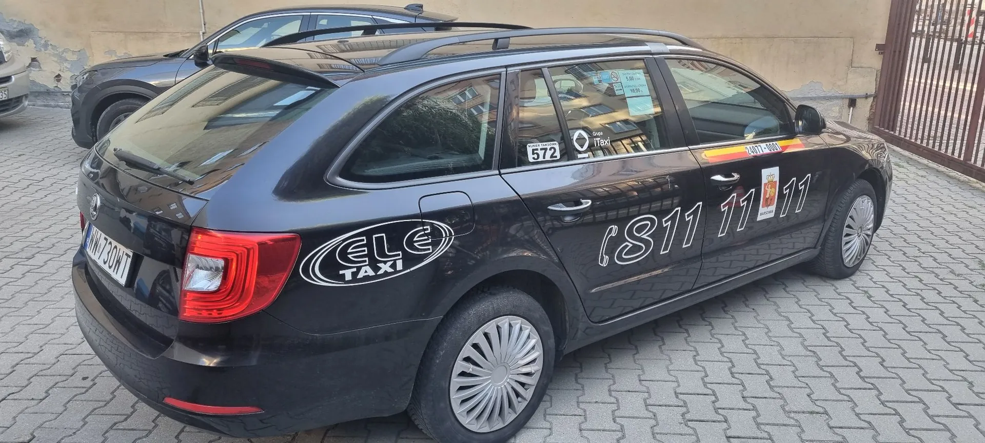 Czarny samochód typu kombi zaparkowany na kostce brukowej. Na boku pojazdu znajdują się białe napisy: logo 'ELE TAXI' na tylnym błotniku oraz numer telefonu '(811 11 11)' na drzwiach. Na szybie bocznej widoczne są naklejki, w tym jedna z numerem '572' oraz napis 'iTaxi'. Samochód ma tablicę rejestracyjną 'WN 730WT'. W tle widoczny jest fragment innego ciemnego samochodu oraz jasna ściana budynku.