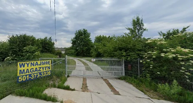 Widok na wjazd na teren ogrodzony metalowym płotem i bramą. Po lewej stronie znajduje się żółta tablica z niebieskim napisem: 'WYNAJMIE MAGAZYNY 501-373-3326'. Droga prowadząca za bramę jest betonowa, z widocznymi śladami kół. W tle oraz po bokach widoczne są gęste zielone krzewy i drzewa pod zachmurzonym niebem.