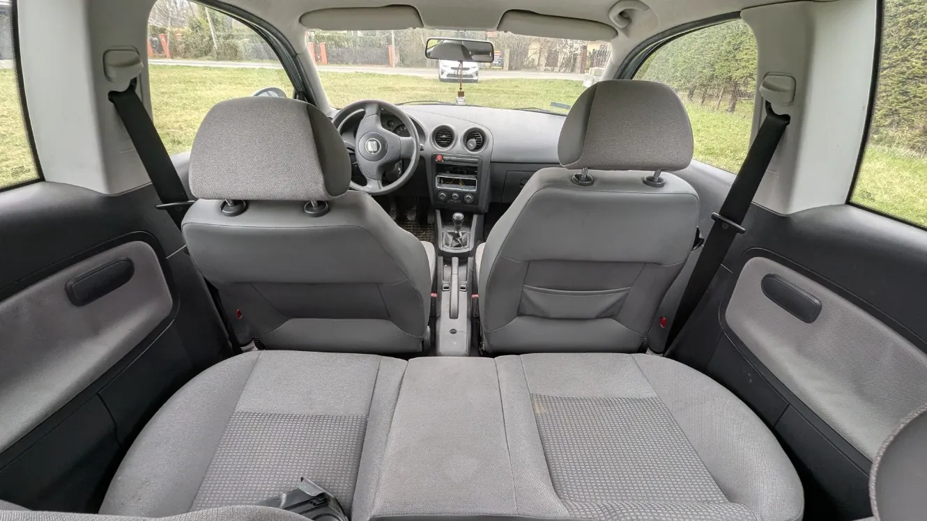 Wnętrze samochodu widziane z tyłu. Szare fotele przednie i tylne z poduszkami pod głowę, kierownica z logo Hondy, deska rozdzielcza z przyciskami i radioodtwarzaczem, dźwignia zmiany biegów w środku. Widoczne pasy bezpieczeństwa, uchwyty drzwiowe oraz zewnętrzny krajobraz z trawą i innym samochodem za przednią szybą. W lusterku wstecznym wiszący przedmiot.