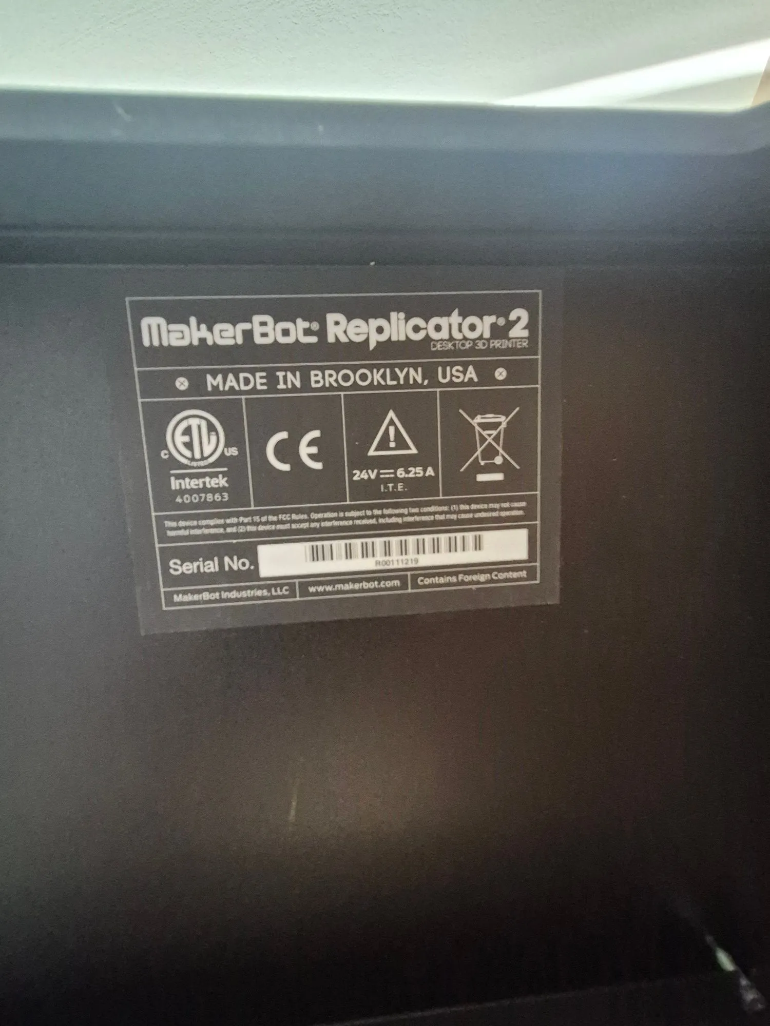 Zdjęcie tabliczki znamionowej drukarki 3D MakerBot Replicator 2. Na tabliczce widnieje napis 'MakerBot Replicator 2 DESKTOP 3D PRINTER' oraz 'MADE IN BROOKLYN, USA'. Widoczne są certyfikaty: znak ETL Intertek (numer 4007863), znak CE oraz symbol ostrzegawczy z trójkątem. Podano parametry zasilania: '24V ⎓ 6.25 A I.T.E.'. Na dole znajduje się pole 'Serial No.' z kodem kreskowym i numerem R00111219, a także nazwa firmy 'MakerBot Industries, LLC', adres strony 'www.makerbot.com' oraz dopisek 'Contains Foreign Content'.