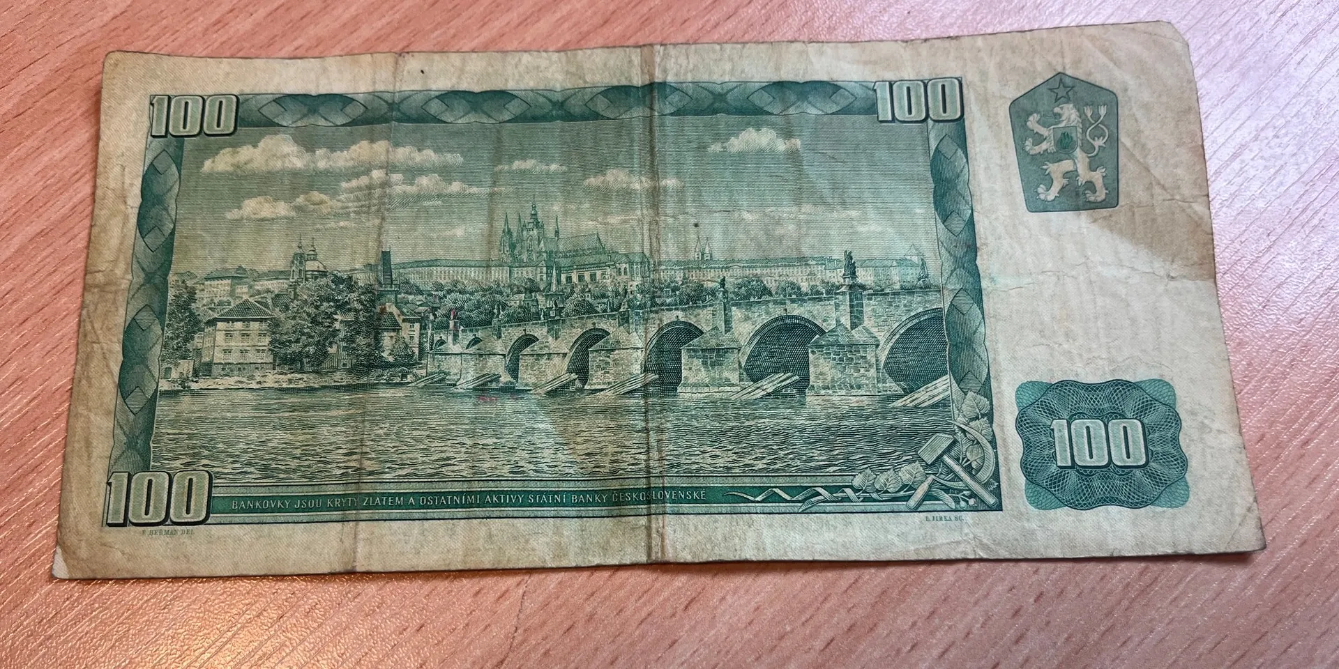 Papierowy banknot o nominale 100, przedstawiający panoramę miasta z mostem i zamkiem w tle. Na banknocie widnieje liczba 100 w kilku miejscach oraz tekst w dolnej części: 'BANKOVKY JSOU KRYTY ZLATEM A OSTATNIMI AKTIVY STATNI BANKY CESKO-SLOVENSKE'. W prawym górnym rogu znajduje się herb z lwem. Banknot jest pognieciony i ma lekko pożółkły kolor.