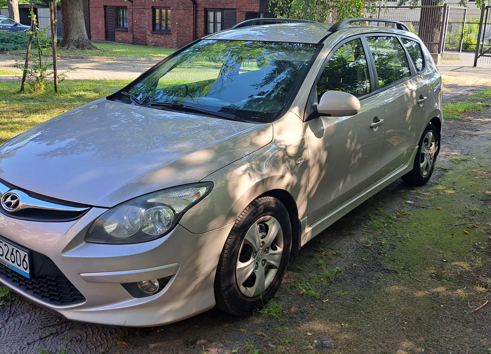 Srebrny samochód marki Hyundai w nadwoziu kombi, zaparkowany na trawiastym podjeździe przed ceglanym budynkiem. Widoczny jest fragment tablicy rejestracyjnej z numerem 52606. Auto posiada relingi dachowe i jest oświetlone promieniami słońca prześwitującymi przez liście drzew.