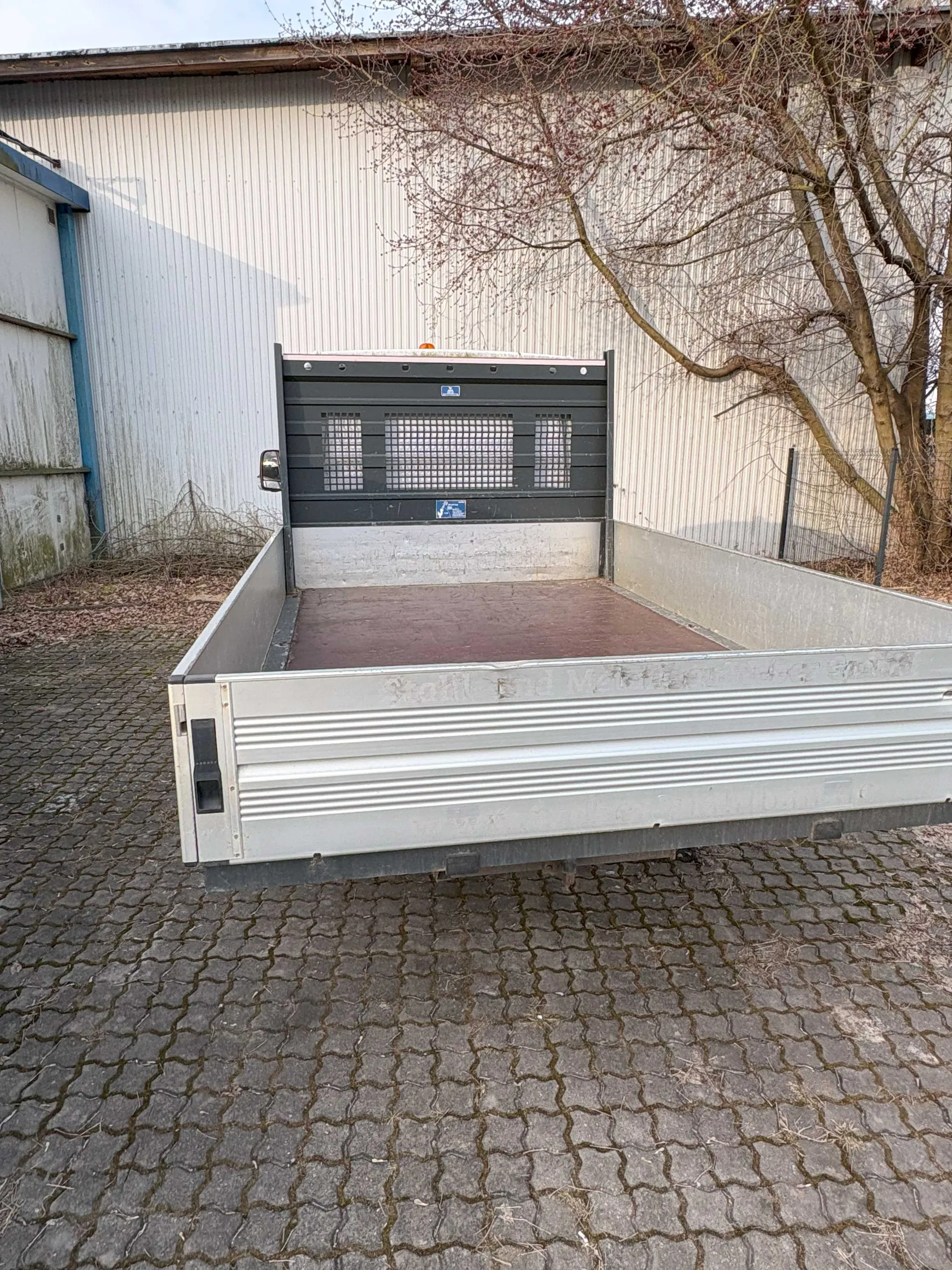 Zdjęcie przedstawia tylną część przyczepy lub platformy ciężarowej z metalowym, srebrnym zbiornikiem o poziomych żeberkach. Wnętrze platformy ma brązowe dno i czarną przód z siatkowymi otworami oraz dwoma niebieskimi naklejkami. Tło stanowi biały, metalowy budynek przemysłowy z pionowymi listwami, po lewej stronie widoczna jest niebiesko-biała brama, a po prawej suche drzewa. Podłoga wykonana z kamiennych, szarych kostek. Na zewnętrznej krawędzi platformy widoczny jest nieczytelny, wypukły napis.