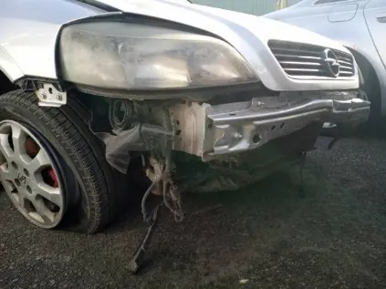 Zdjęcie przedstawia przód białego samochodu marki Mercedes z poważnie uszkodzonym zderzakiem. Widoczne są pęknięte reflektory, zdeformowana maska oraz częściowo zwisający zderzak. W tle znajduje się fragment innego pojazdu. Samochód stoi na nawierzchni asfaltowej.