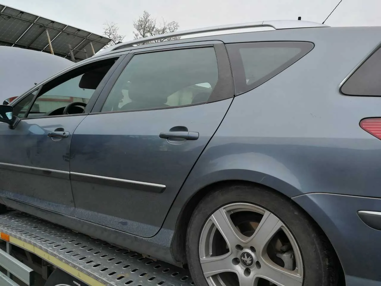 Szary samochód typu kombi marki Peugeot z widocznym logo na felgach, stojący na metalowej platformie z perforowanym blatem. W tle budowla z panelami słonecznymi i drzewa bez liści. Niebo jest pochmurne. Samochód posiada dachówkę i srebrne klamki drzwi.