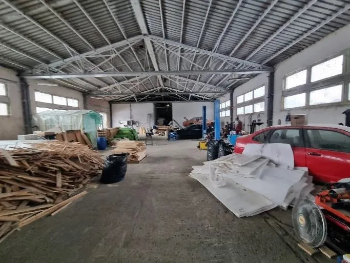 Wnętrze przemysłowej hali z metalowym dachem i belkami. Po lewej stronie stosy drewnianych desek i czarne worki na śmieci. W centrum betonowa podłoga, po prawej czerwony samochód osobowy oraz białe panele i materiały. W tle widoczne są maszyny, niebieska kolumna oraz okna po obu stronach hali. Oświetlenie naturalne wpada przez okna.