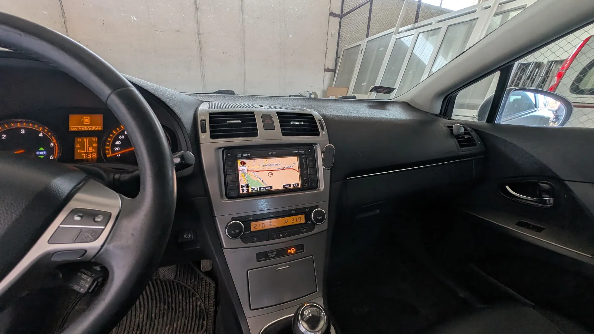 Widok z wnętrza samochodu z perspektywy kierowcy. Widoczna jest czarna kierownica po lewej stronie, deska rozdzielcza z licznikiem prędkości, ekranem nawigacji GPS w konsoli centralnej oraz panelem sterowania klimatyzacją z cyfrowymi wyświetlaczami temperatury 21.0. W tle widać fragmenty siedzeń oraz lusterko boczne odbijające otoczenie.