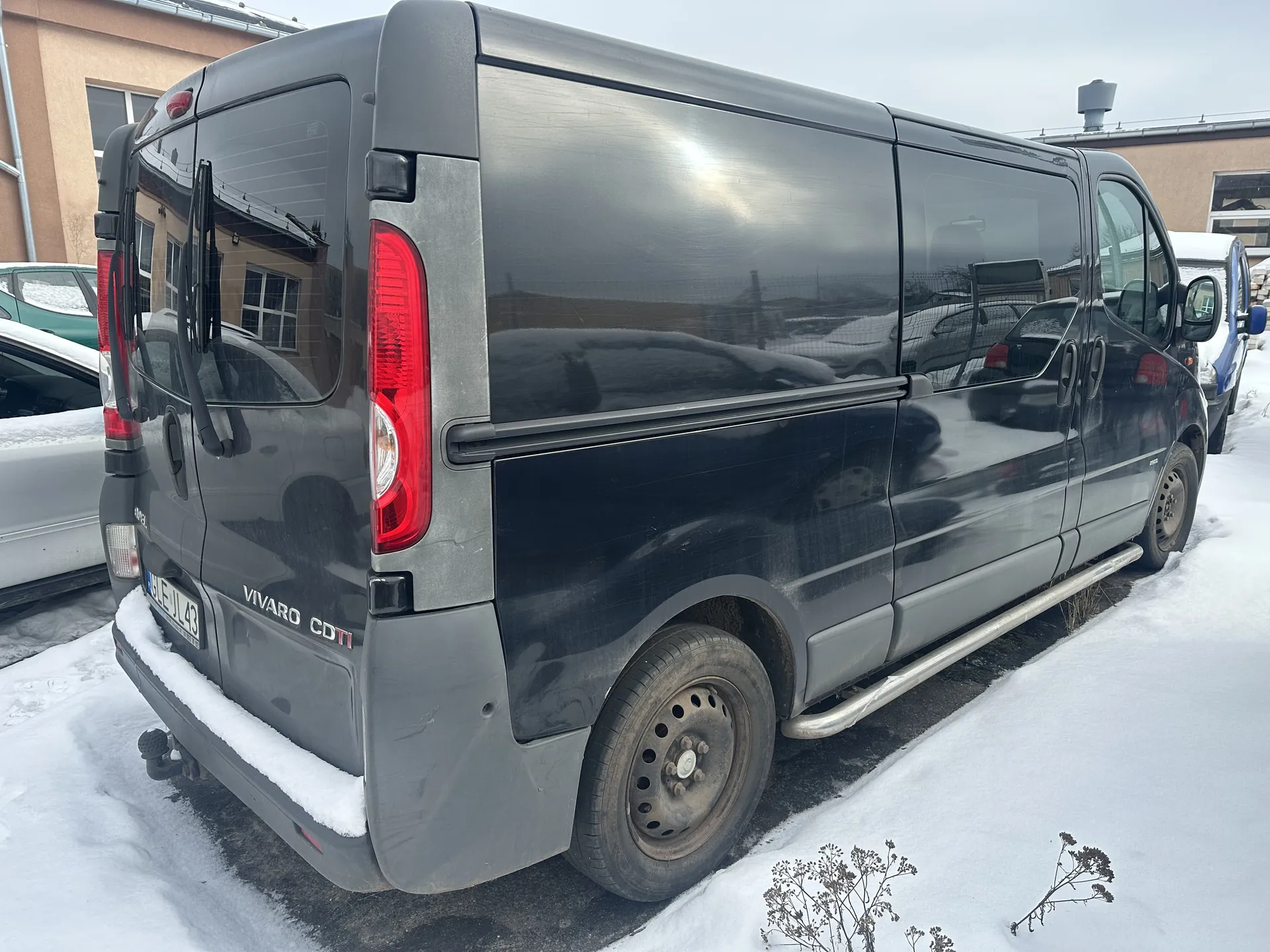 Ciemnoszary samochód dostawczy Opel Vivaro CDTI zaparkowany na śnieżnej powierzchni. Widoczny tył pojazdu z czerwonymi światłami, tablicą rejestracyjną EJL 43 oraz napisem VIVARO CDTI na bagażniku. Pojazd otoczony jest innymi samochodami, w tle budynki o beżowych ścianach i oknach. Na ziemi leży śnieg, a na niebie panuje pochmurna pogoda.