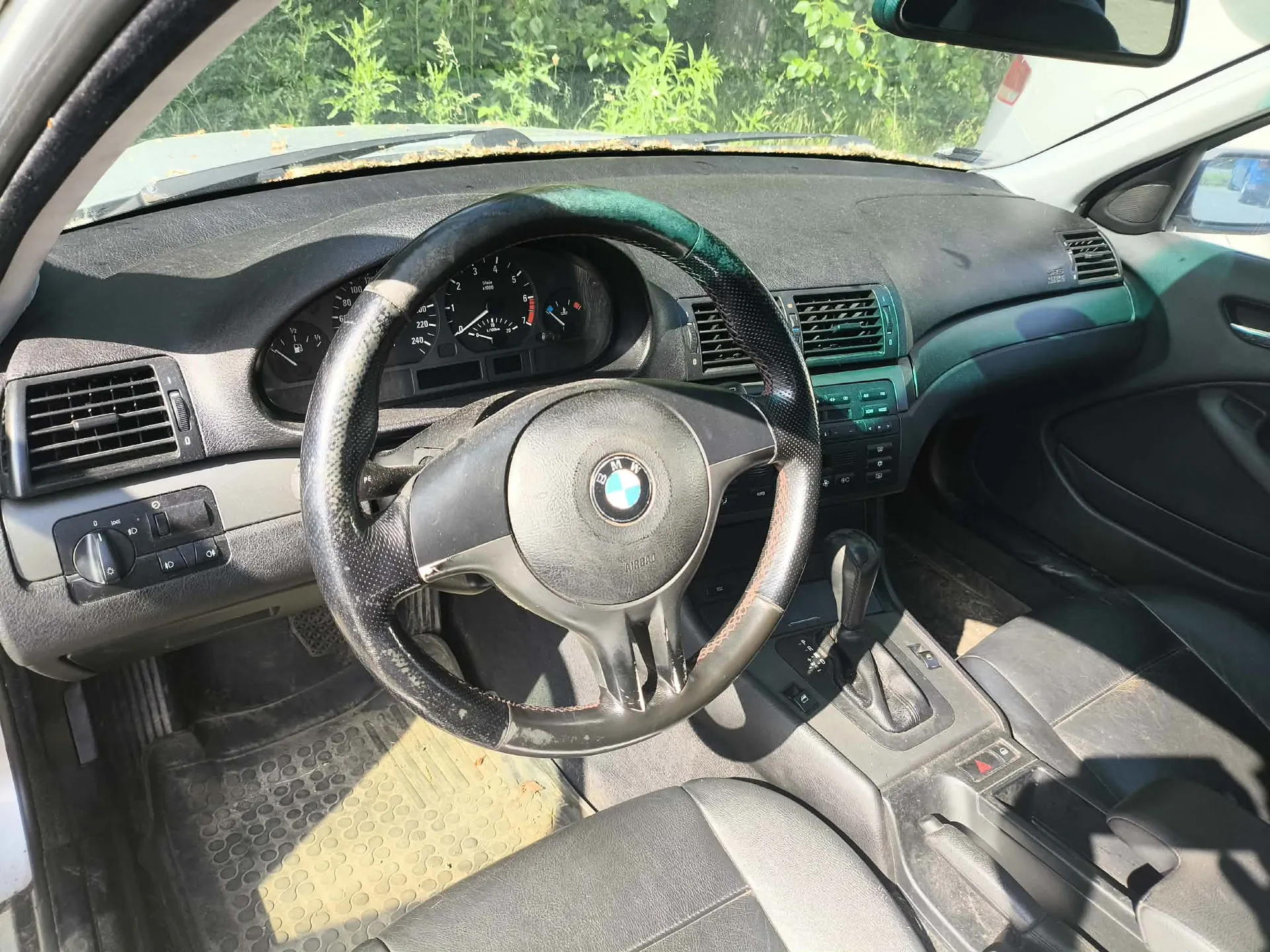 Wnętrze samochodu marki BMW z perspektywy kierowcy. Widoczna jest czarna skórzana kierownica z logo BMW w centrum, deska rozdzielcza z zegarami, kratki nawiewów oraz lewarek zmiany biegów w konsoli środkowej. Podłoga wyłożona jest gumowymi dywanikami. Przez przednią szybę widoczna jest zieleń roślinności.