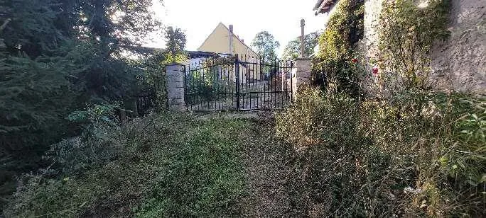 Zdjęcie przedstawia żółty dom z trójkątnym dachem, czarną metalową bramę między kamiennymi filarami, porośniętą trawą i krzewami przestrzeń na pierwszym planie, drzewa po lewej stronie oraz kamienną ścianę z pnącza po prawej. W tle widoczne są dodatkowe drzewa i jasne niebo.