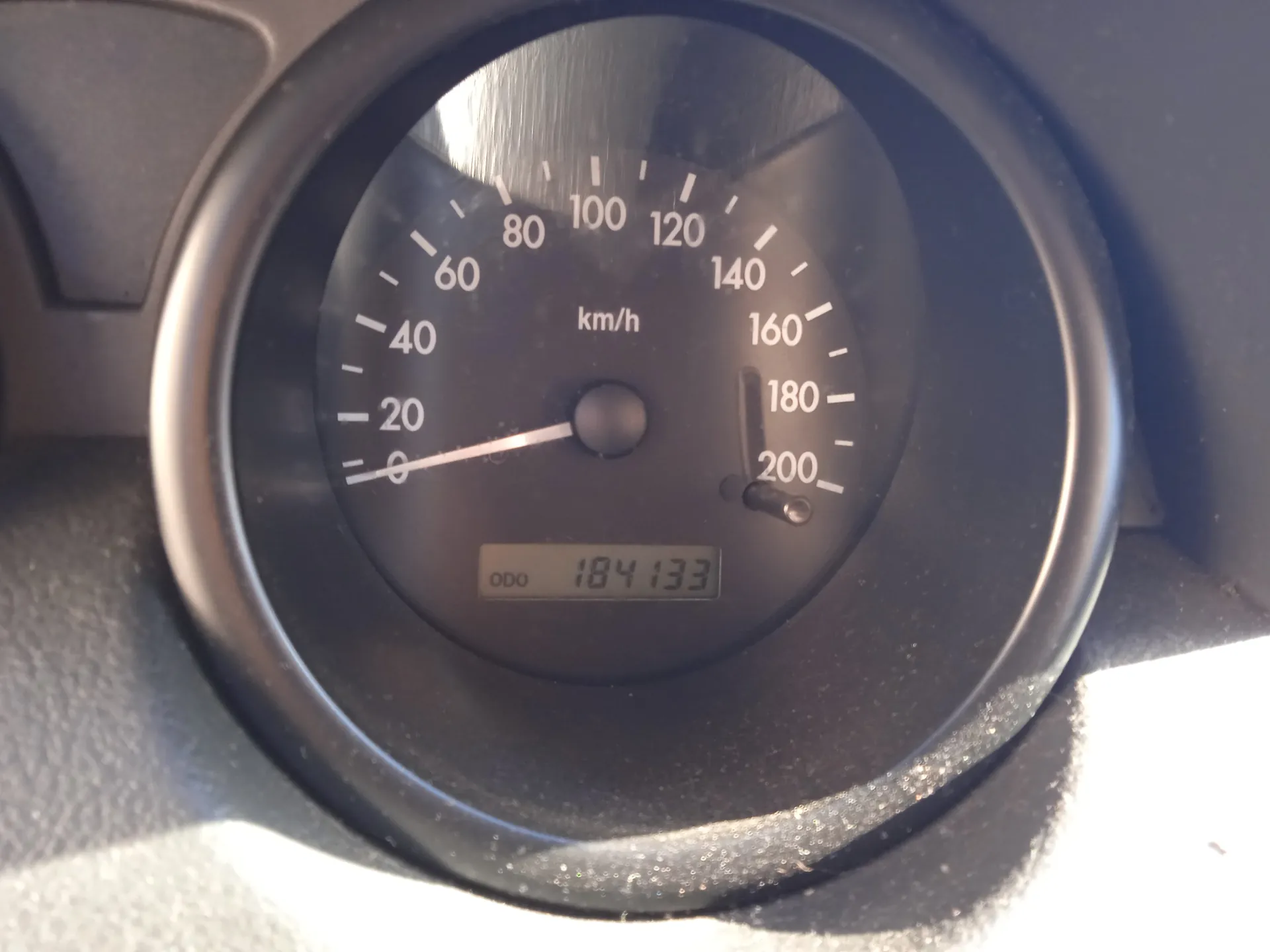 Zbliżenie na czarny samochodowy wskaźnik prędkości z białymi znacznikami i cyframi od 0 do 200 km/h. Wskazówka znajduje się na pozycji 0. W centrum widnieje napis 'km/h'. Poniżej cyfrowy licznik przebiegu z napisem 'ODO' i wartością '184133'. Tło stanowi fragment ciemnoszarej deski rozdzielczej.