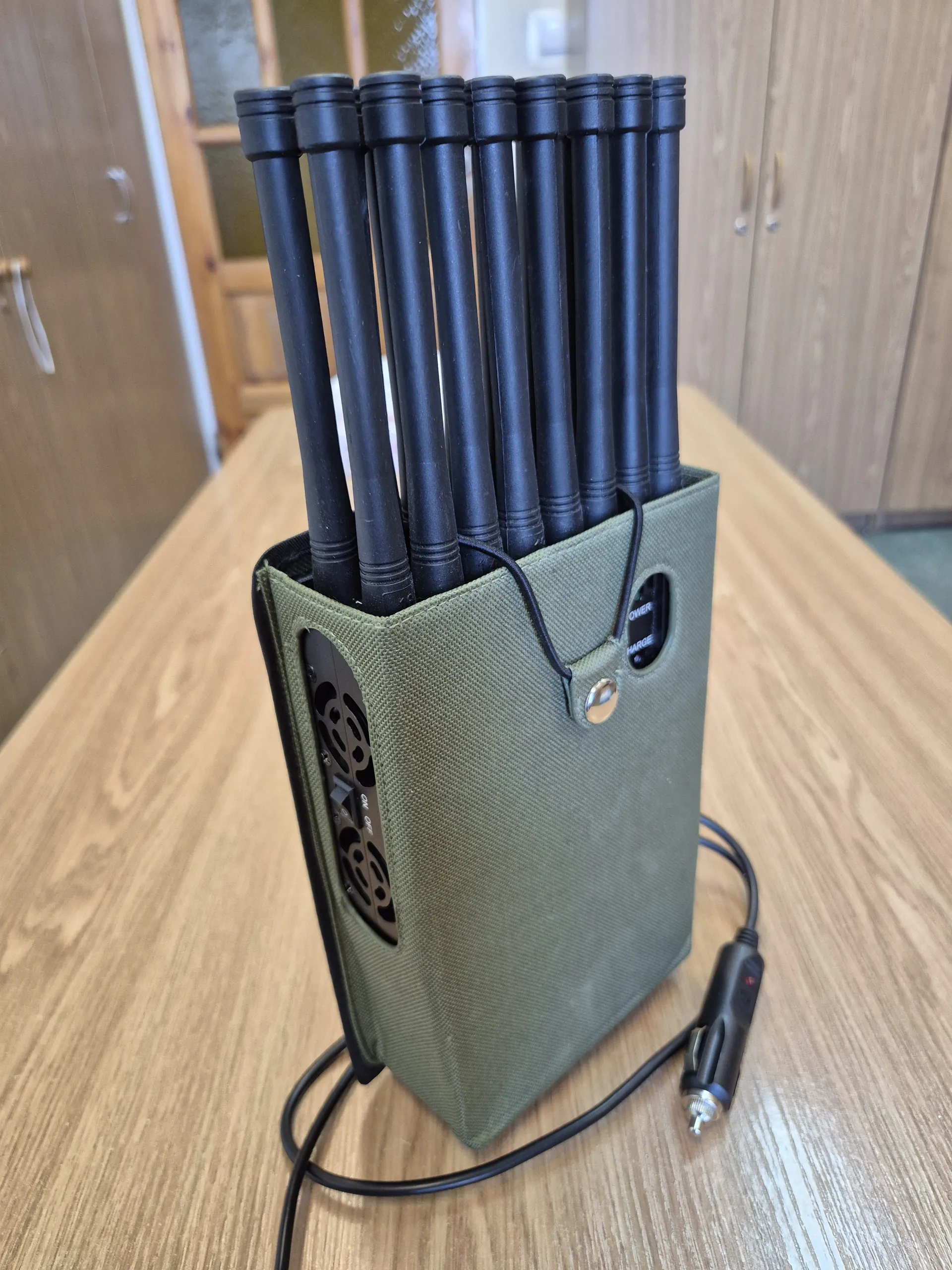 Zielona tkaninowa torebka z czarnymi cylindrycznymi elementami wystającymi z jednej strony. Do torebki podłączony jest czarny kabel z zapalniczkowym wtyczkiem samochodowym. Na torebce widoczne są przyciski, otwory wentylacyjne oraz napisy 'ON' i 'OFF'. Tło stanowi drewniany stół i szafki z drewnianymi uchwytami.