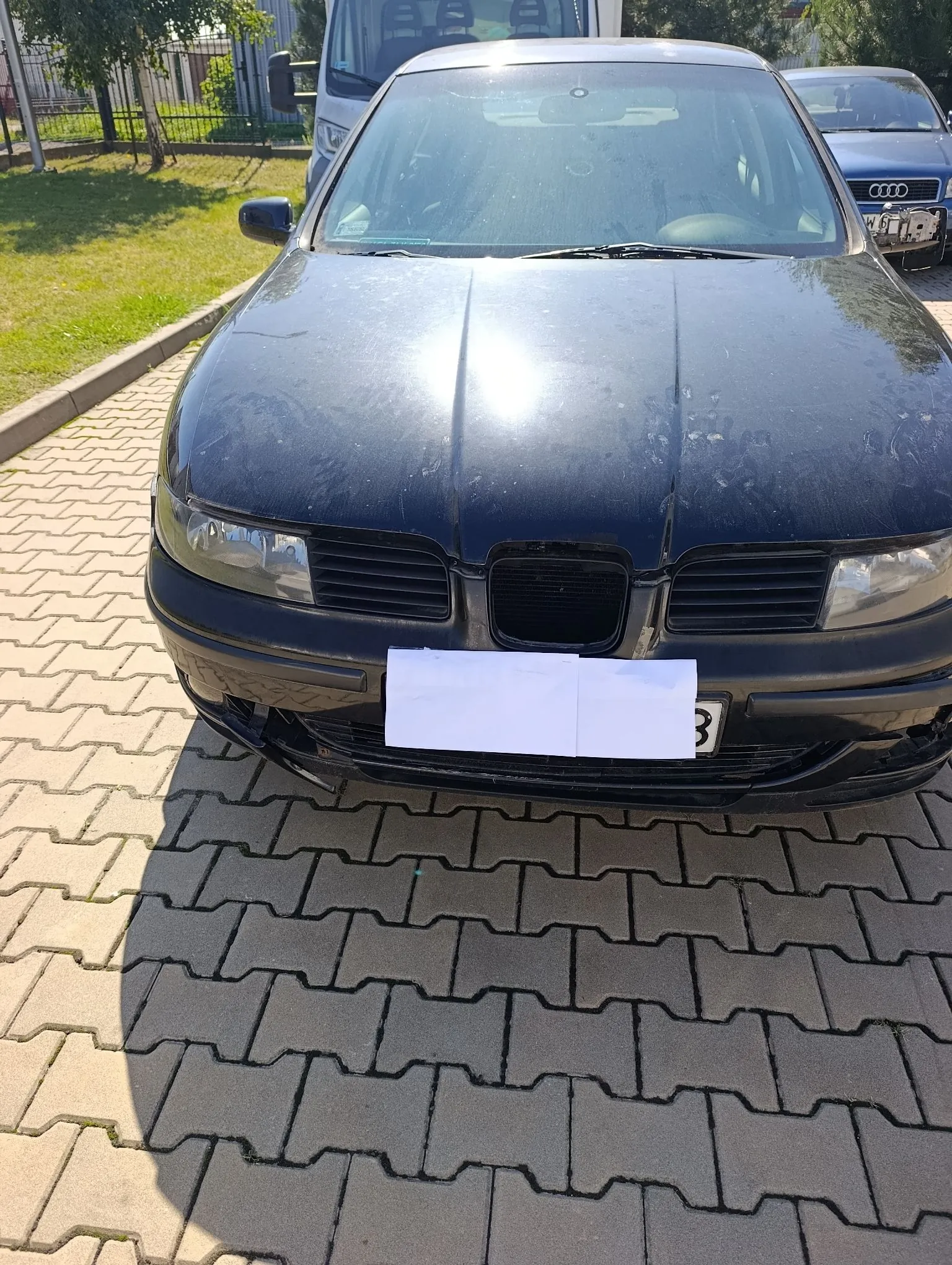 Zdjęcie przodu czarnego samochodu osobowego zaparkowanego na kostce brukowej. Samochód ma zakryty numer rejestracyjny białym prostokątem. W tle widoczny jest fragment białego vana oraz srebrnego samochodu marki Audi, a także trawnik i drzewa.