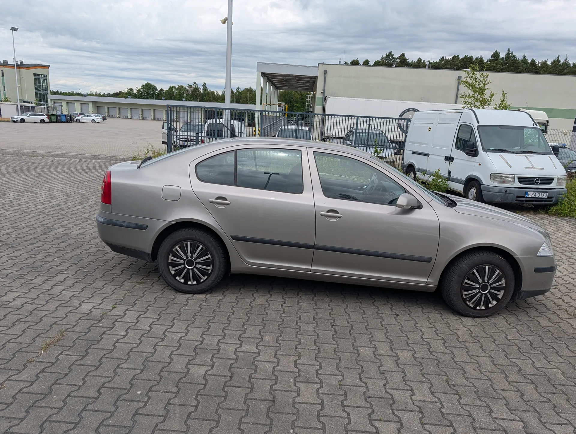 Samochód osobowy srebrny sedan zaparkowany na brukowanym placu. Obok biały furgon z tablicą rejestracyjną FZA 31X3. W tle przemysłowe budynki, ogrodzenie metalowe, inne pojazdy, drzewa i zachmurzone niebo.