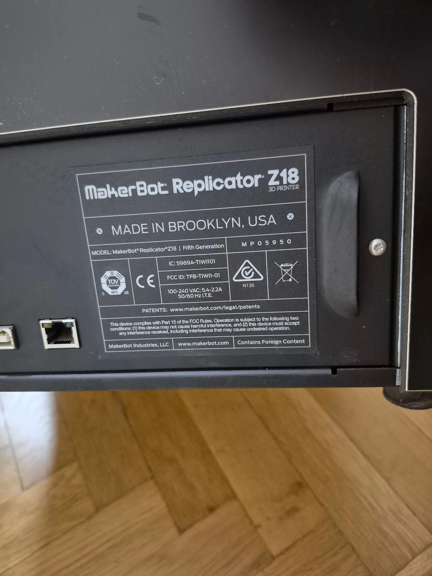 Zdjęcie tabliczki znamionowej drukarki 3D MakerBot Replicator Z18. Na czarnym tle znajduje się biała etykieta z tekstem: 'MakerBot Replicator Z18 3D PRINTER', 'MADE IN BROOKLYN, USA'. Widoczne są dane techniczne: MODEL: MakerBot Replicator Z18 | Fifth Generation, numer seryjny MP05950, IC: 5969A-TIW1101, FCC ID: TFB-TIW11-01, napięcie 100-240 VAC; 5A-2.2A 50/60 Hz 1.1E. Na etykiecie znajdują się również certyfikaty TÜV SÜD, CE oraz symbol ostrzegawczy. Na dole widnieje adres strony www.makerbot.com/legal/patents oraz informacja 'MakerBot Industries, LLC' i 'Contains Foreign Content'. Po lewej stronie widoczny jest port sieciowy Ethernet.