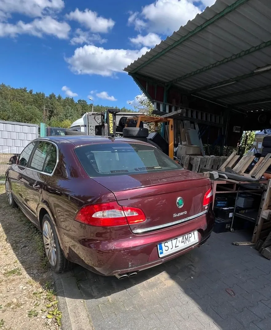 Bordowy samochód osobowy marki Skoda Superb zaparkowany tyłem na kostce brukowej. W tle widoczny jest zadaszony obiekt gospodarczy, wózek widłowy marki Hyster oraz las pod niebem z białymi chmurami. Tablica rejestracyjna samochodu to SJZ 4MP1.