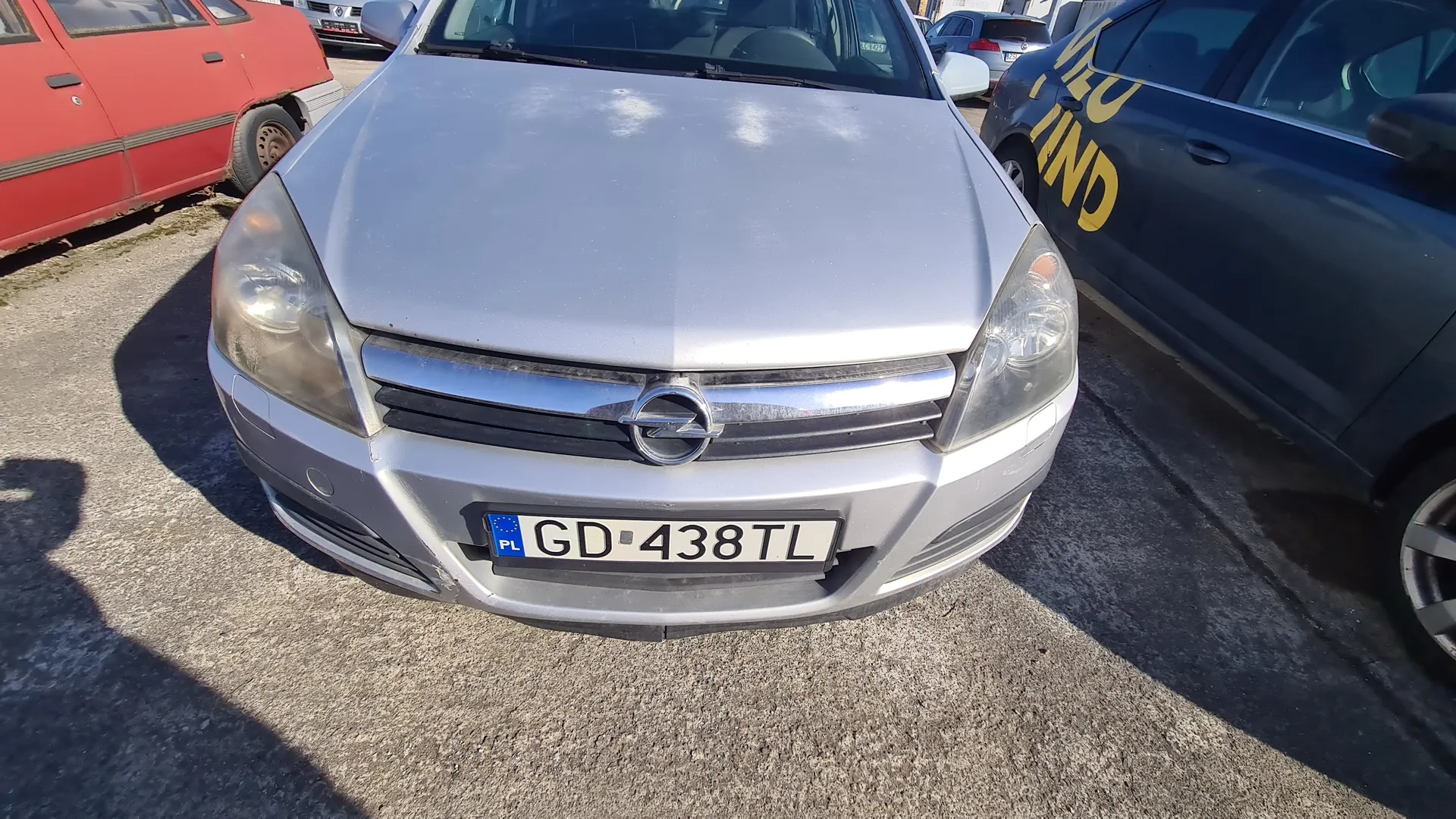 Przedni widok srebrnego samochodu marki Opel z tablicą rejestracyjną GD-438TL (PL). Po lewej stronie czerwony samochód, po prawej ciemnoniebieski samochód z żółtym napisem MEU LAND. Tło stanowi asfaltowy parking z innymi pojazdami w oddali.