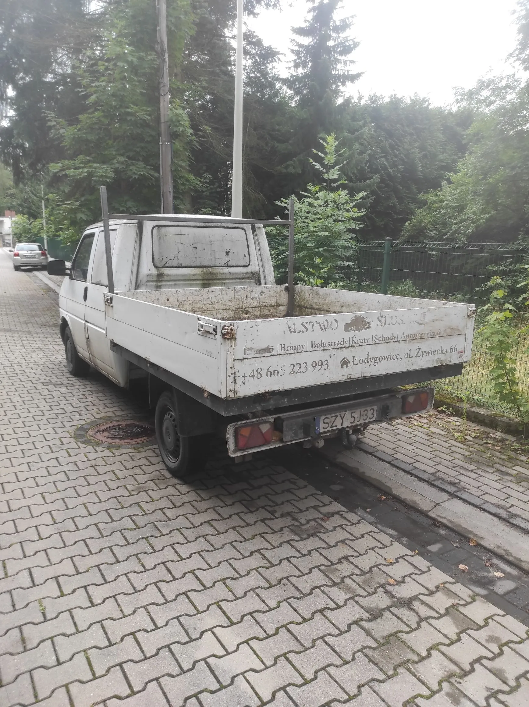 Biały, stary samochód dostawczy typu pick-up z otwartą paką, zaparkowany na kostce brukowej. Pojazd jest w złym stanie technicznym, z widocznymi śladami rdzy i zużycia. W tle znajduje się gęsta roślinność, drzewa iglaste i liściaste oraz metalowy płot.