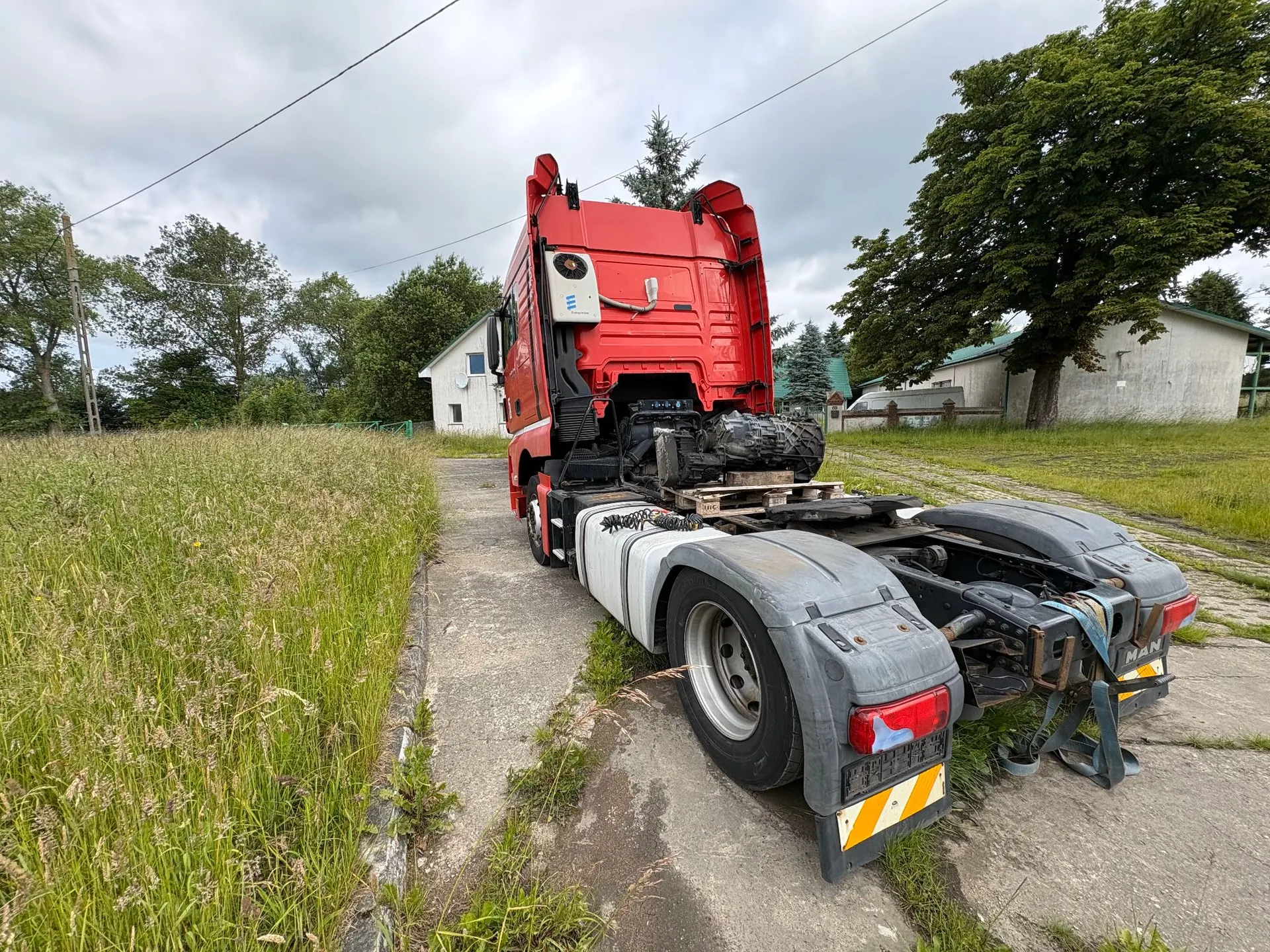 Czerwona ciągnika siodłowego zaparkowana na betonowej drodze obok trawiastego pola. Pojazd jest widoczny od tyłu, z odsłoniętym podwoziem i ramą. W tle widać zielone drzewa, krzewy oraz biały budynek mieszkalny pod zachmurzonym niebem.