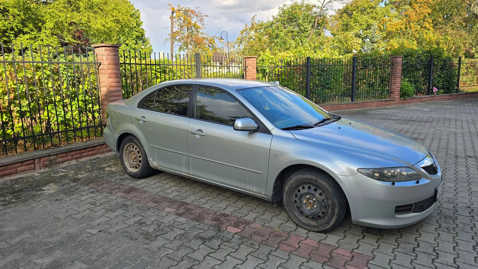 Srebrny samochód osobowy typu sedan zaparkowany na kostce brukowej. Pojazd stoi bokiem do obiektywu, w tle znajduje się czarny metalowy płot z ceglanymi słupkami oraz zielone drzewa i krzewy.