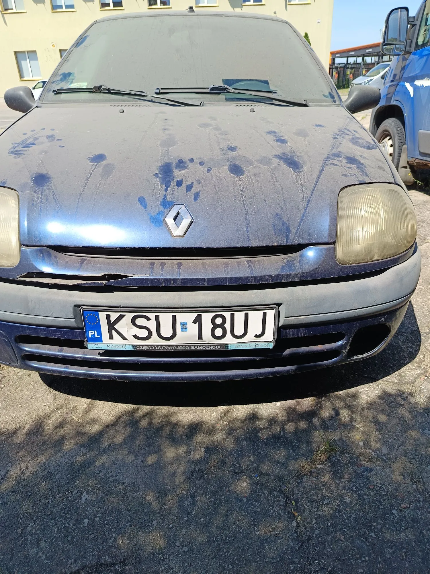 Zdjęcie przodu niebieskiego samochodu marki Renault. Na masce widoczne są liczne zacieki i plamy. Samochód posiada polską tablicę rejestracyjną o numerze KSU 18UJ. Reflektory są zmatowiałe. W tle widoczny jest fragment żółtego budynku oraz fragment niebieskiej ciężarówki po prawej stronie.