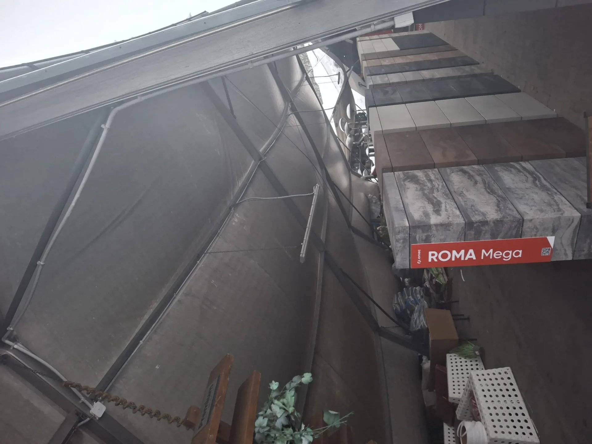 Widok pod daszkiem z materiałów budowlanych. W centrum czerwony pionowy znak z białym tekstem 'ROMA Mega' oraz mniejszym napisem 'fajniec' na górze. Po lewej stronie białe kratowane pojemniki, brązowe meble i zielone rośliny. Po prawej stronie stosy płytek w kolorach szarym, brązowym i beżowym. Daszek z szarym materiałem, widoczne rury i oświetlenie. Tło z niebieskim niebem i dodatkowymi konstrukcjami.
