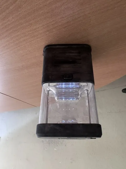 Czarna latarka kempingowa z przezroczystym, plastikowym kloszem, leżąca poziomo na brązowym blacie z drewna.