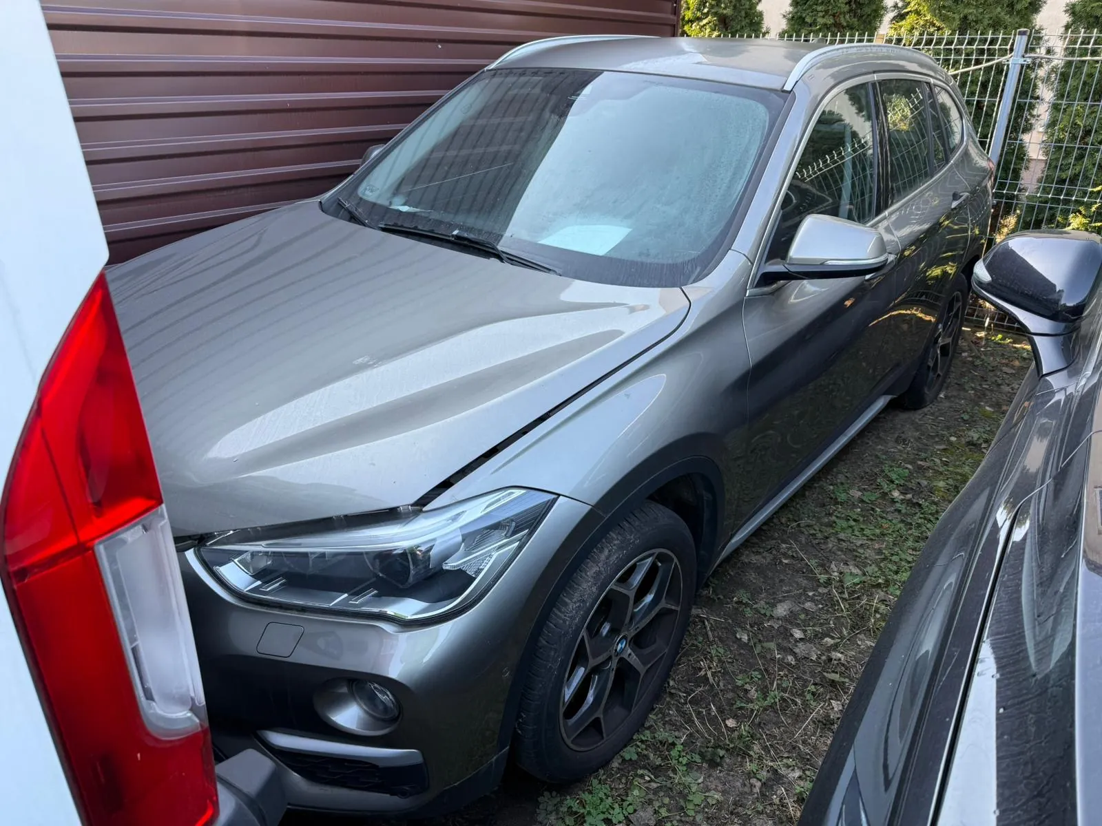 Srebrny SUV zaparkowany między dwoma innymi pojazdami. Po lewej stronie widoczny czerwony tylny reflektor białego samochodu, po prawej czarny boczny lusterko innego pojazdu. W tle brązowa brama garażowa, metalowe ogrodzenie i zielone krzewy. Podłoga pokryta suchą trawą i ziemią. Samochód ma czarne felgi i czystą karoserię.