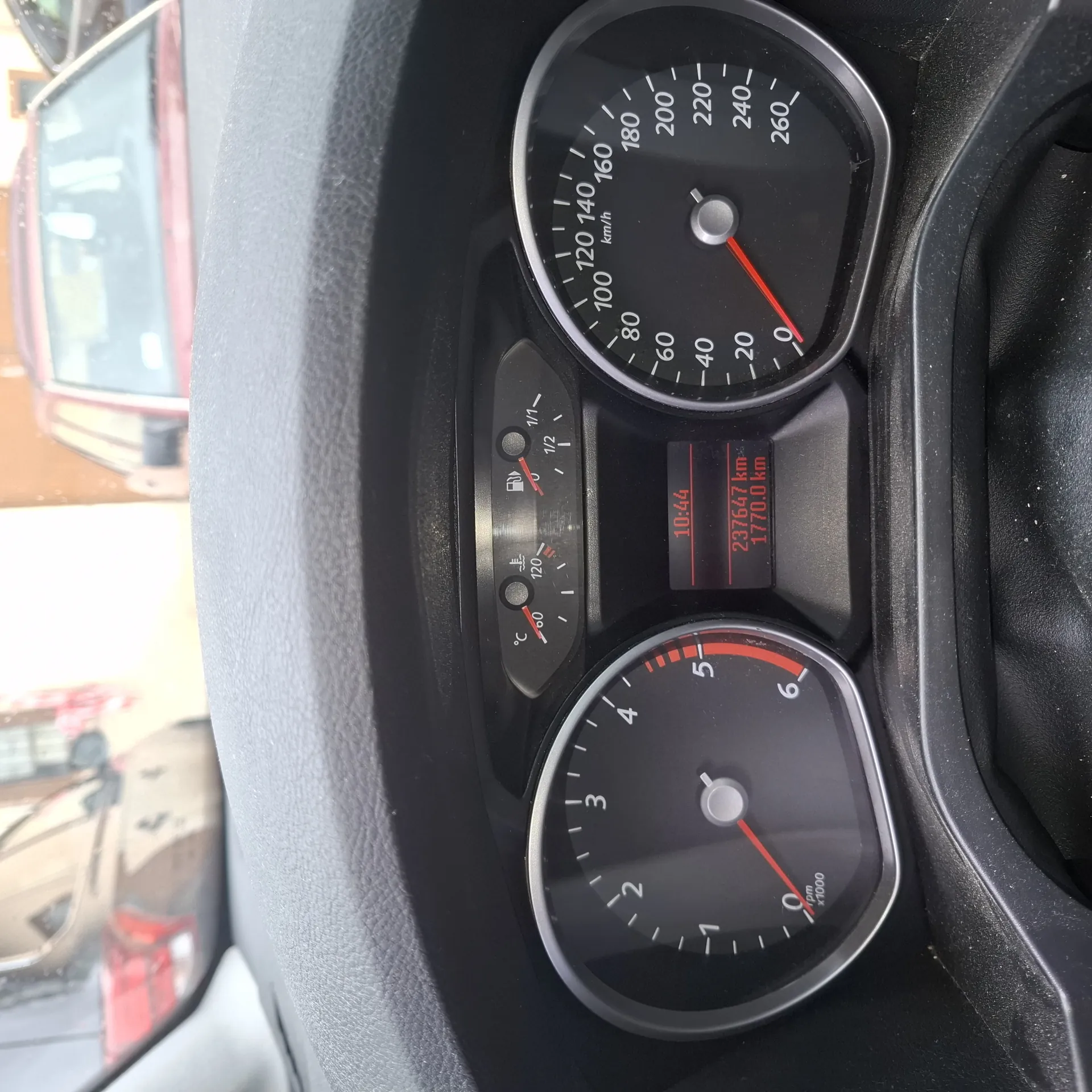 Zbliżenie na deski rozdzielcze samochodu. W górnej części znajduje się prędkościomierz z zakresem do 260 km/h, wskazówka na 0. Poniżej zegar obrotomierz z zakresem do 6 (x1000 obr./min), wskazówka na 0. Po lewej stronie pionowy wskaźnik paliwa (połowa zbiornika) i temperatury chłodniczki (ok. 60°C). W centralnej części wyświetlacz cyfrowy z czerwonymi cyframi: godzina 10:44, przebieg 237647 km, licznik dzienny 1770.0 km. W tle widoczne fragmenty lusterka bocznego i ulicy.