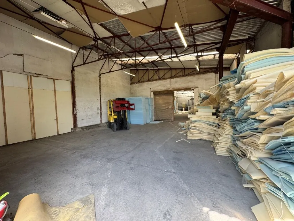 Wnętrze dużego, industrialnego magazynu lub hali z betonową podłogą. Po prawej stronie znajduje się wysoki stos dużych arkuszy pianki w kolorach białym, kremowym i błękitnym. Po lewej stronie widoczna jest jasna ściana z panelami. W głębi pomieszczenia stoi żółty wózek widłowy oraz błękitne bloki materiału. Sufit jest konstrukcją stalową z widocznymi uszkodzeniami i zwisającymi fragmentami izolacji oraz lampami fluorescencyjnymi.