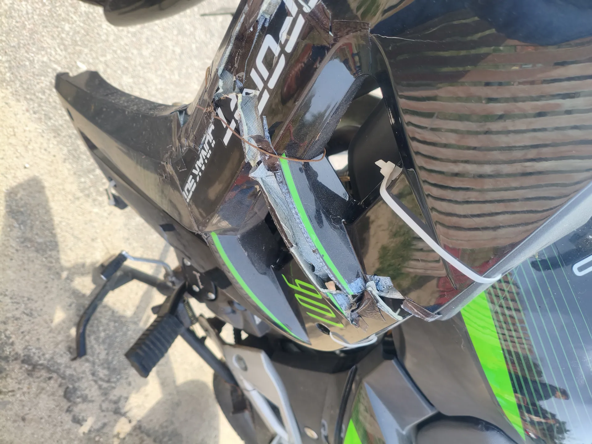 Zbliżenie na uszkodzoną czarną osłonę przednią motocykla z zielonymi akcentami. Widoczne są pęknięcia i taśma klejąca naprawiająca uszkodzenia. Na osłonie zielonymi cyframi widnieje numer '901', a białymi literami fragmenty napisów 'SPORT' oraz 'JUNAK 50'. W tle asfaltowa powierzchnia oraz odbicie drewnianego ogrodzenia i osób na błyszczącej powierzchni motocykla.