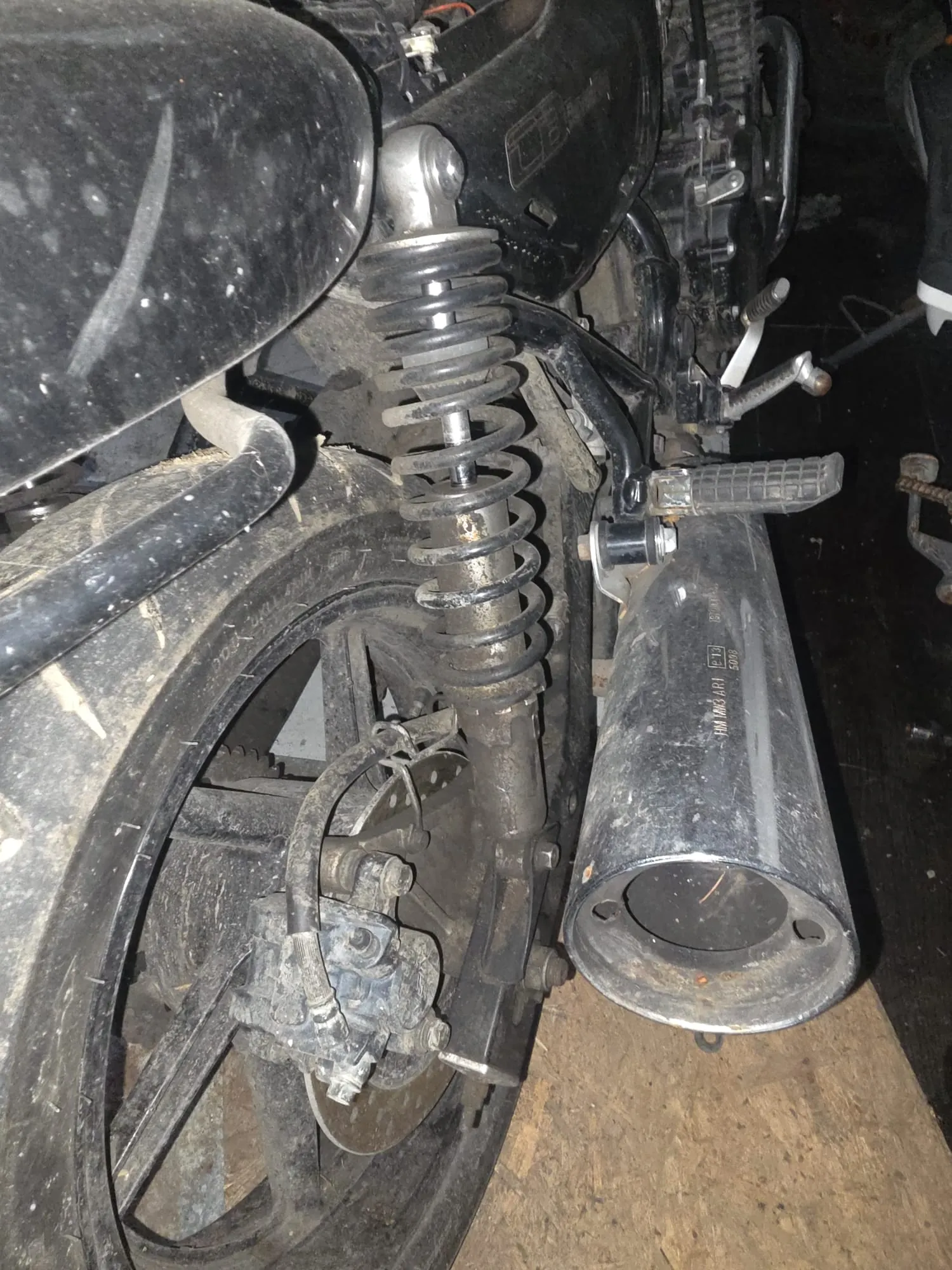 Zbliżenie na tylną część motocykla, obejmujące oponę z kołem, sprężynę amortyzatora, układ hamulcowy oraz wydech. Elementy są zabrudzone, widoczne ślady zużycia. Tło ciemne, prawdopodobnie garaż.
