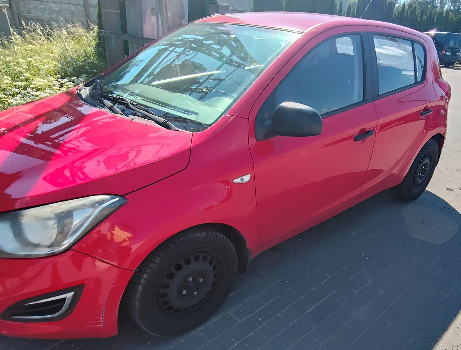 Czerwony samochód osobowy typu hatchback zaparkowany na kostce brukowej. Zdjęcie przedstawia przód i prawy bok pojazdu pod kątem. Samochód ma czarne felgi stalowe i czarne lusterka boczne.