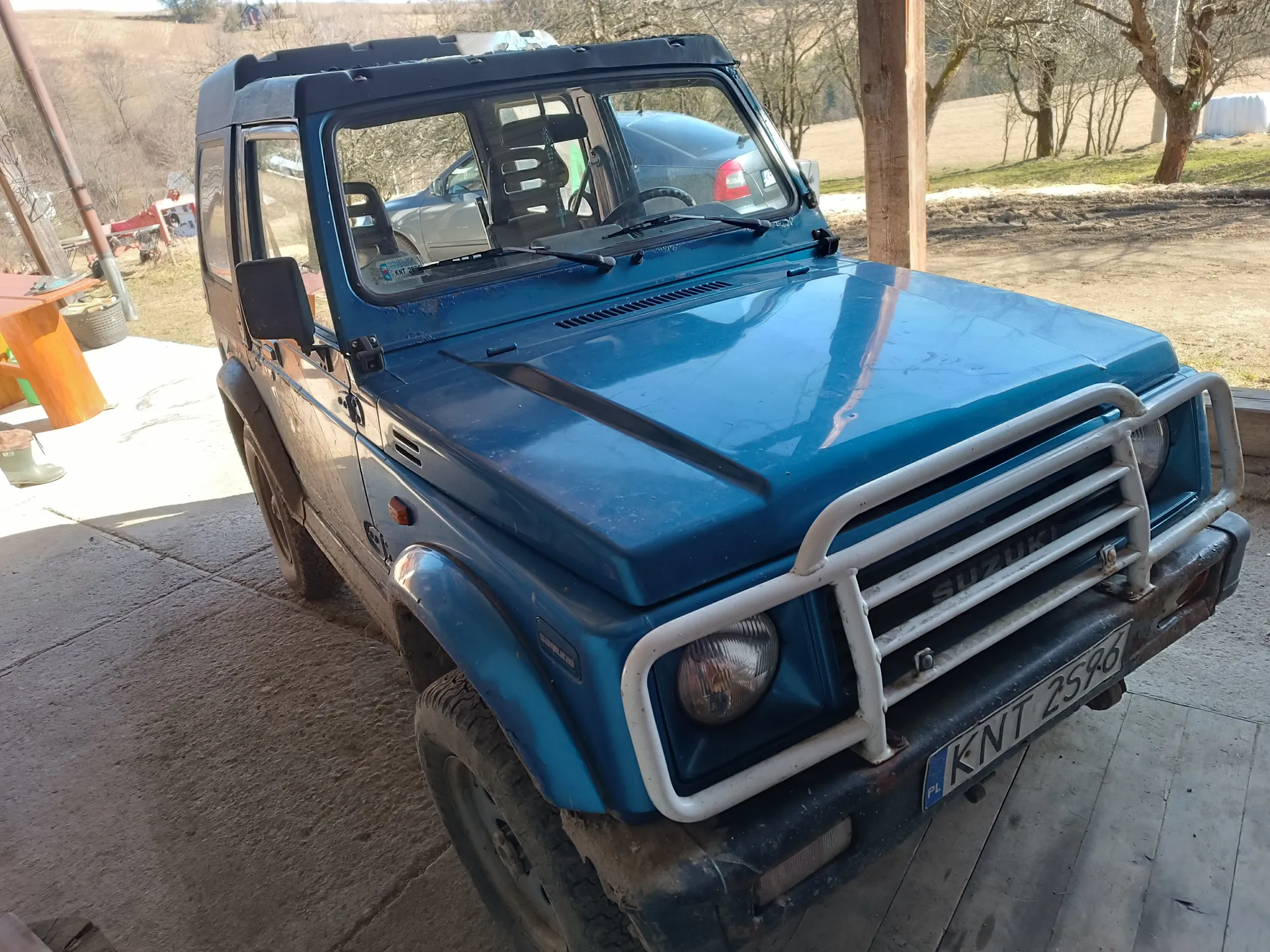 Niebieski samochód terenowy marki Suzuki z białym zderzakiem przednim i tablicą rejestracyjną KNT 2596 (PL). Pojazd stoi na betonowej powierzchni, w tle widoczne są inne samochody, pola, drzewa bez liści oraz drewniana konstrukcja. Na przedniej szybie odbija się srebrny samochód. W tle po lewej stronie znajdują się pomarańczowe elementy i wiadro.