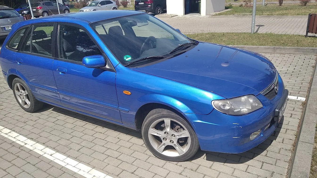 Niebieski samochód osobowy typu kombi zaparkowany na kostce brukowej. Pojazd jest ustawiony pod kątem, widoczny z przodu i z boku. W tle widać inne zaparkowane samochody, fragment białego budynku oraz trawnik.
