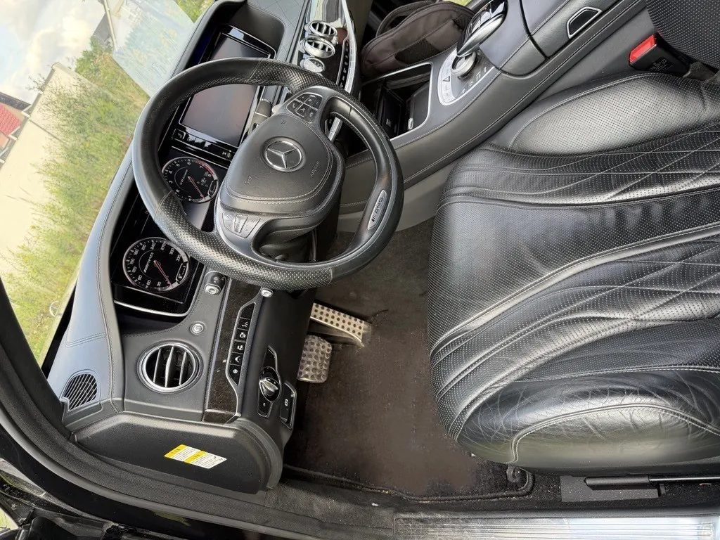 Wnętrze samochodu marki Mercedes-Benz z perspektywy kierowcy. Widoczna jest czarna, skórzana kierownica z logo marki na środku, deska rozdzielcza z zegarami, otwory wentylacyjne oraz czarny, perforowany skórzany fotel. W stopach widoczne są aluminiowe pedały.