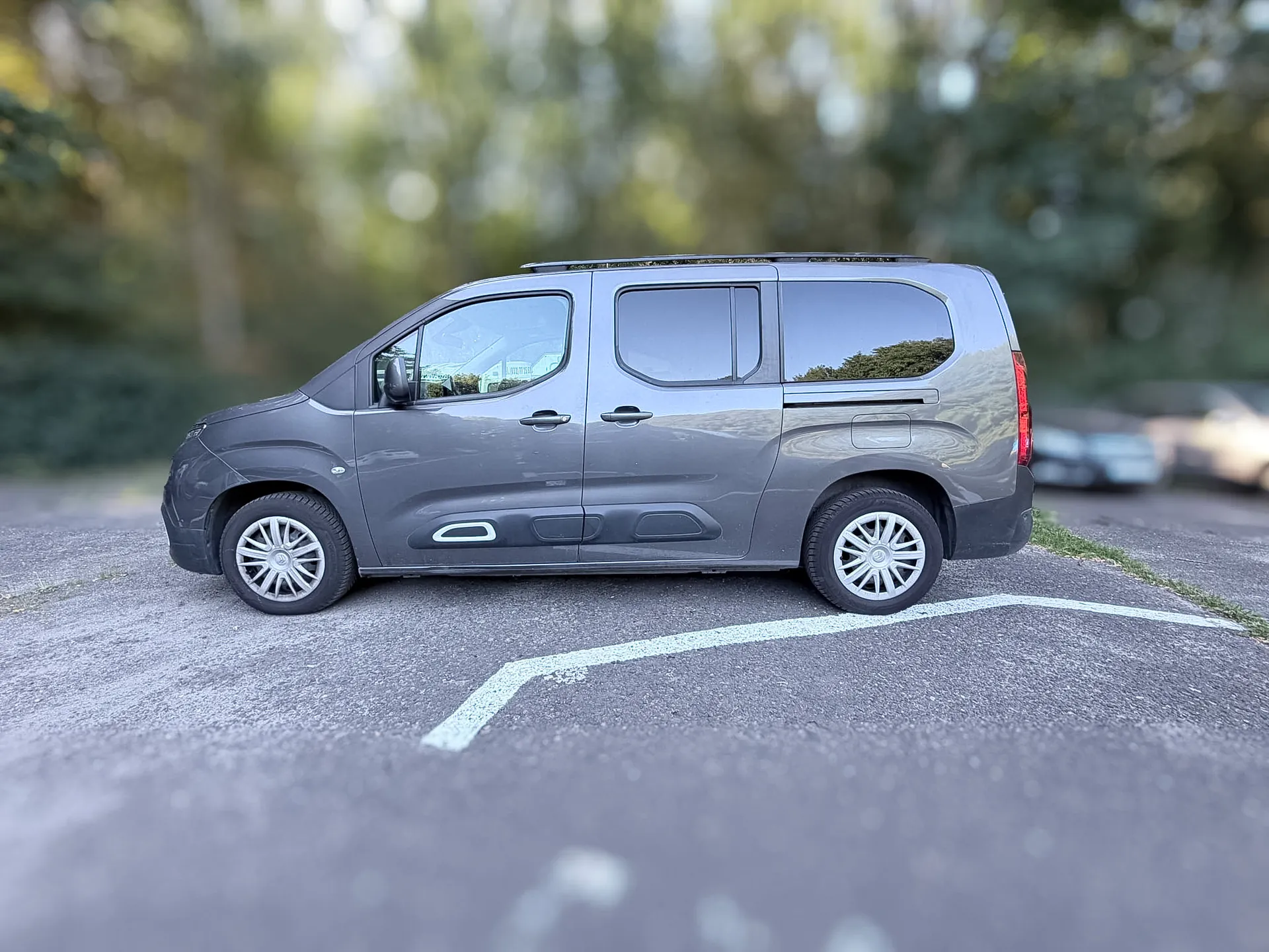 Szary samochód dostawczy typu van zaparkowany bokiem na asfaltowym parkingu. Pojazd ma srebrne felgi aluminiowe i ciemne szyby w tylnej części. W tle widoczne są rozmyte zielone drzewa.