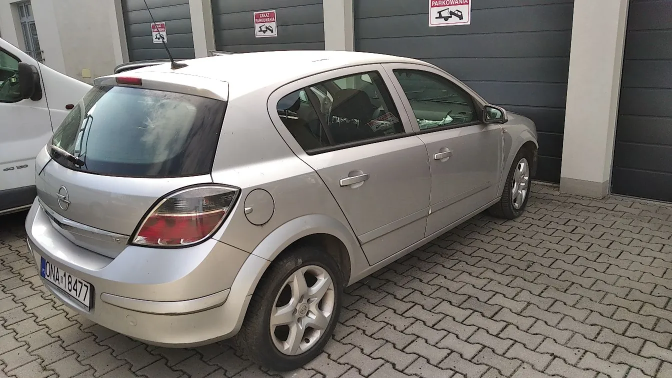 Srebrny samochód marki Opel Astra w nadwoziu hatchback, zaparkowany tyłem do widza na kostce brukowej przed garażami. Na tylnej klapie widoczna jest tablica rejestracyjna o numerze ONA 18477. W tle znajdują się szare drzwi garażowe z tabliczkami informującymi o zakazie parkowania.