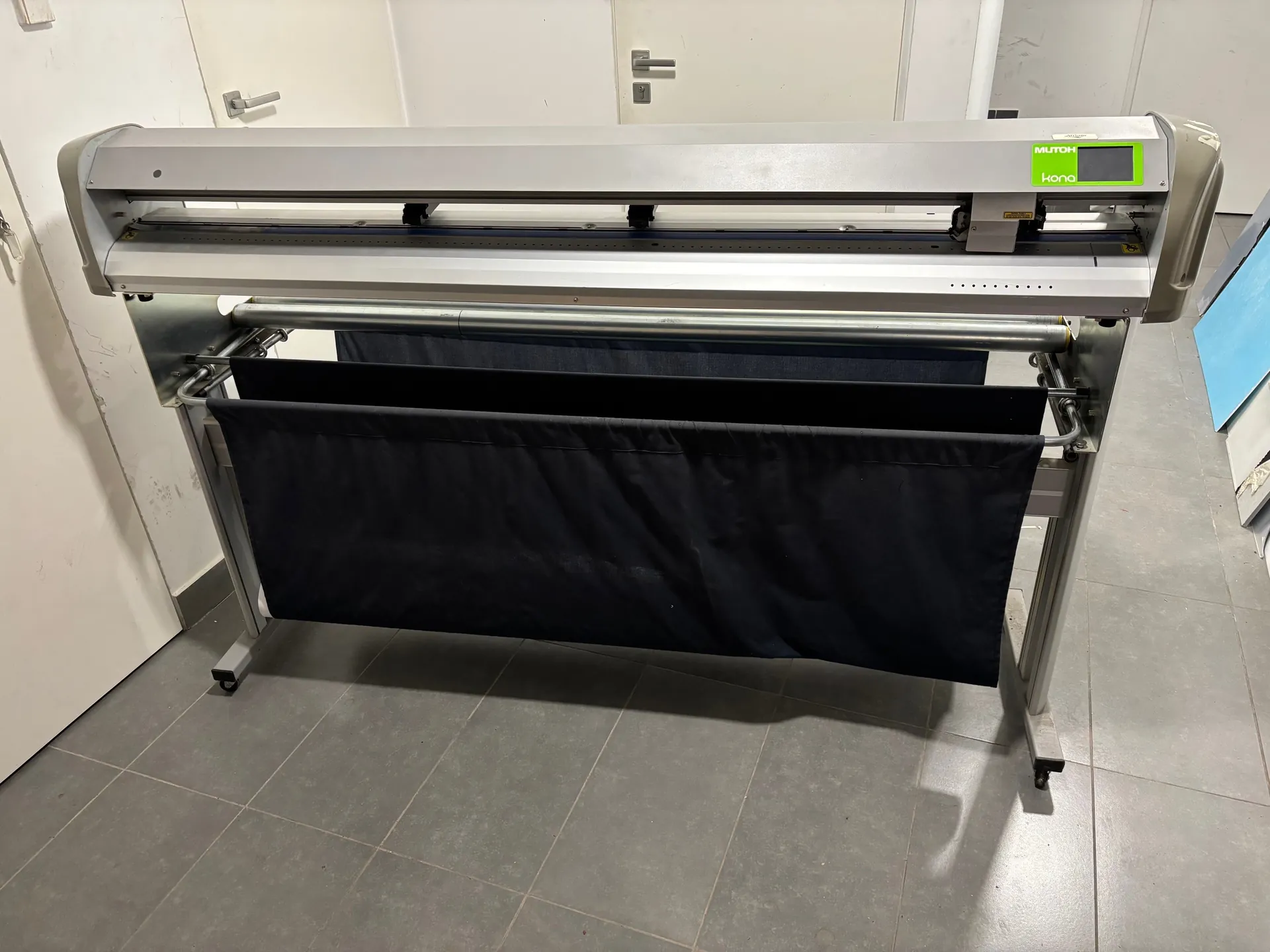 Duży, srebrny ploter lub tnąca maszyna marki MUTOH kona z zieloną etykietą z napisem 'MUTOH kona' na prawym końcu. Maszyna posiada metalowe wały i czarne materiały tekstylne zawieszone na dolnej części. W tle białe drzwi z metalowymi klamkami oraz szare płytki podłogowe. Całość znajduje się w pomieszczeniu o jasnych ścianach.