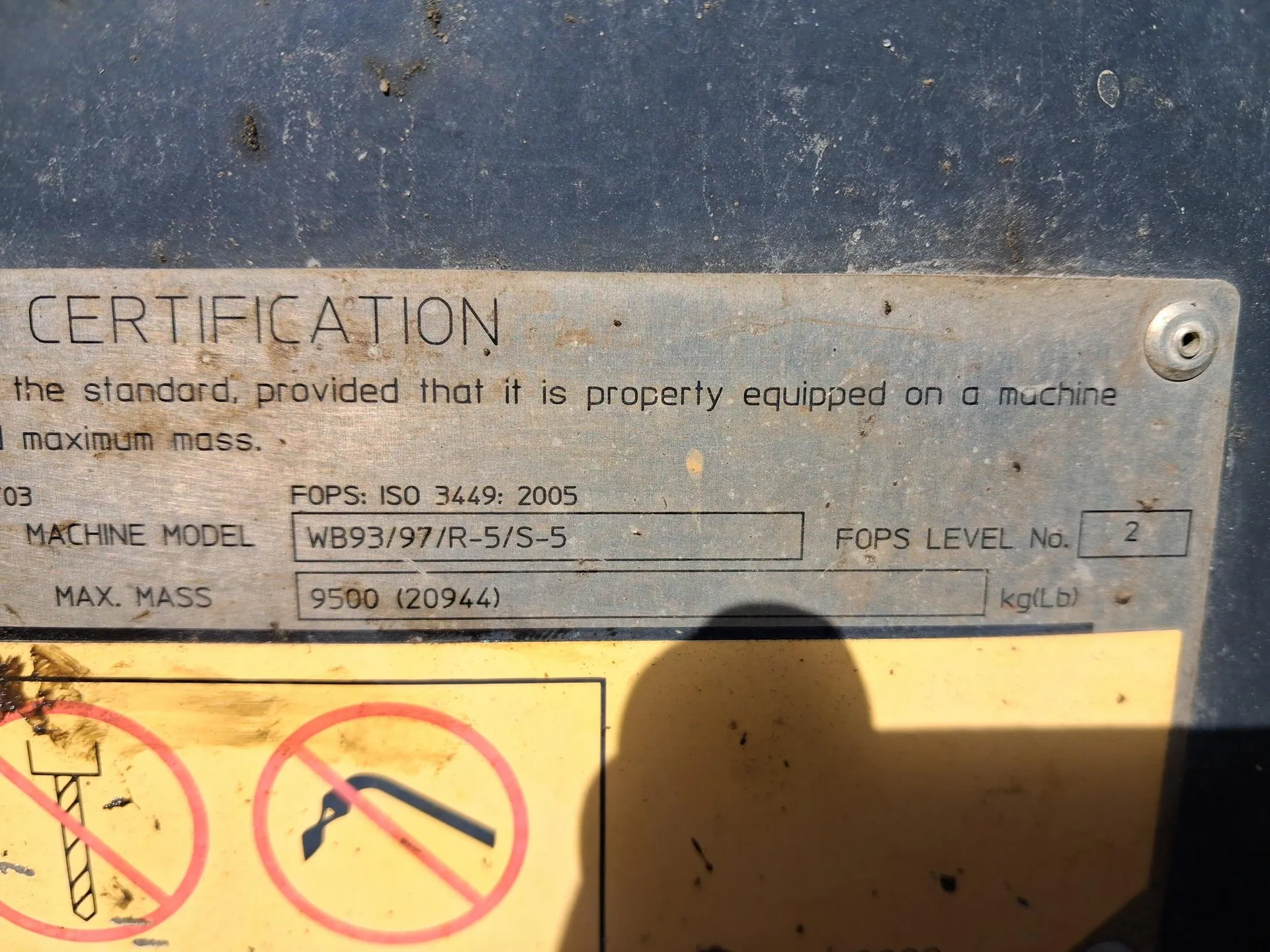 Metalowa tablica z napisem 'CERTIFICATION' na górze. Poniżej widoczny tekst: 'the standard, provided that it is property equipped on a machine maximum mass.' oraz dane techniczne: 'FOPS: ISO 3449: 2005', 'MACHINE MODEL WB93/97/R-5/S-5', 'FOPS LEVEL No. 2', 'MAX. MASS 9500 (20944) kg(Lb)'. Na dole żółta tablica ostrzegawcza z dwoma zakazami w czerwonych kołach: pierwszy przedstawia zakaz używania dźwigu, drugi zakaz używania narzędzia przypominającego łom. Tło stanowi niebieska, zabrudzona powierzchnia metalowa.